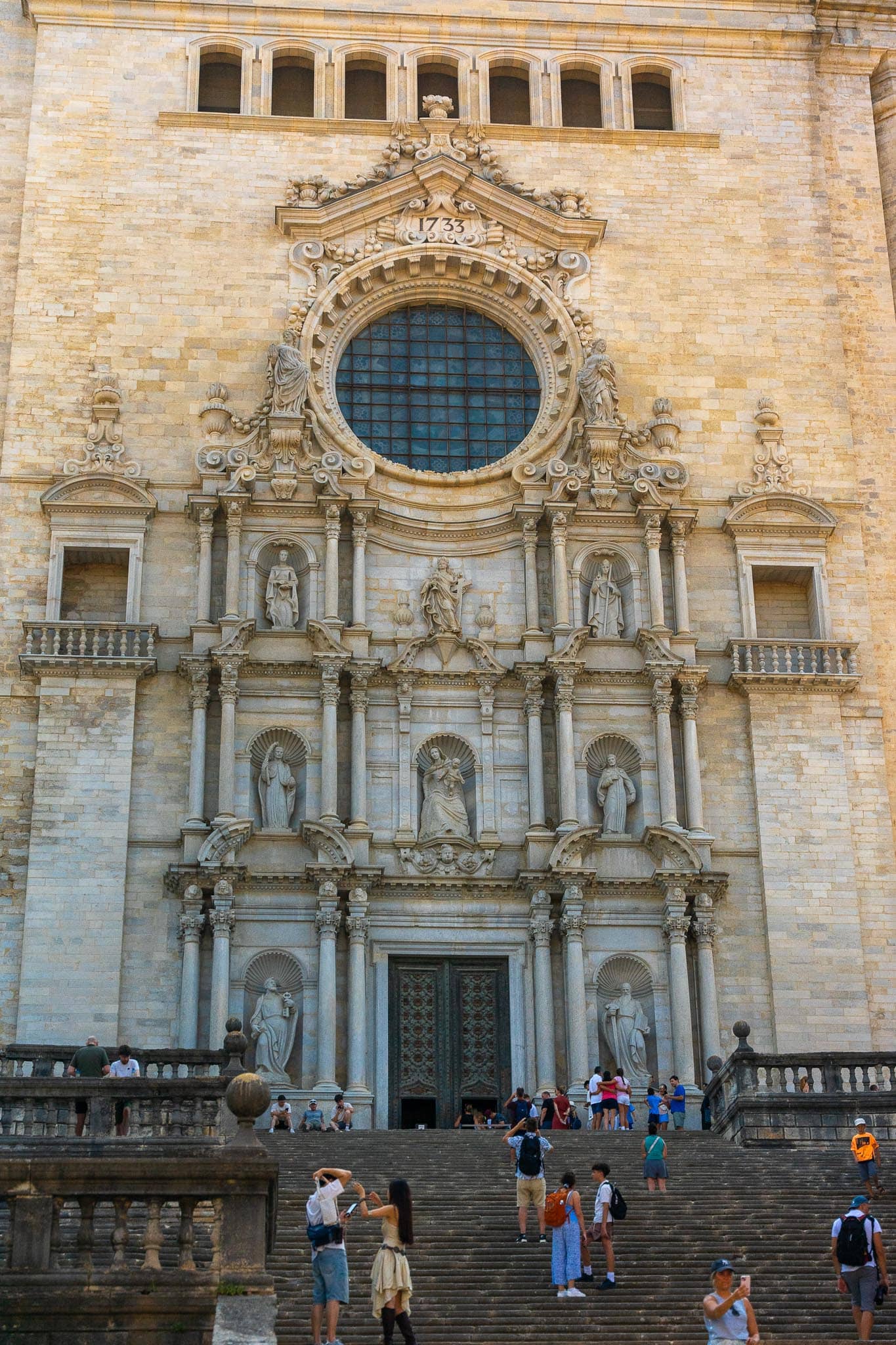 The baroque facade and rose window of Girona cathedral above its long flight of stone steps, with people climbing and sitting on them.