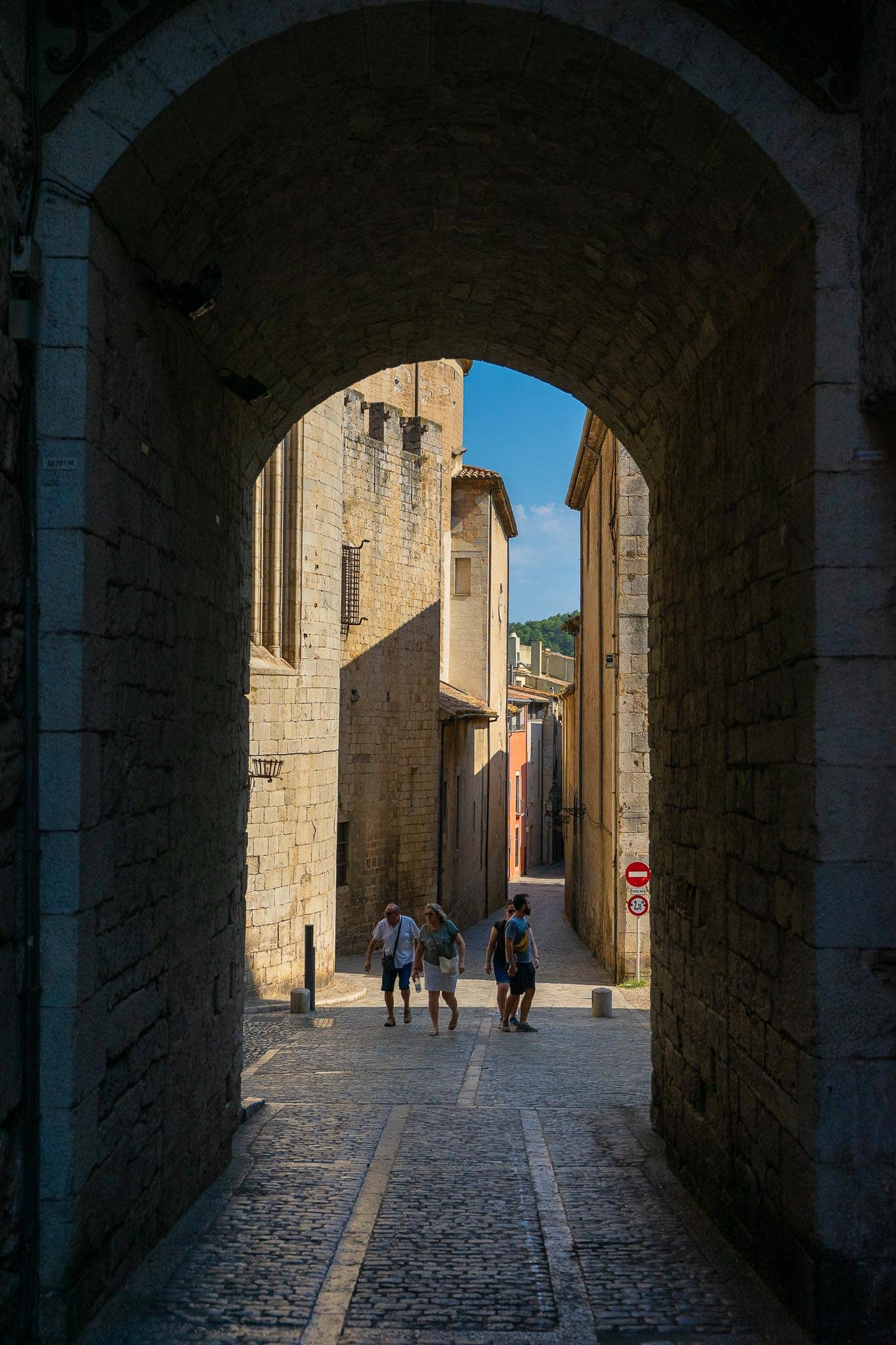 Looking through a deep stone gateway in Girona's old town into a bright narrow lane beyond, with figures walking in the sunlight.