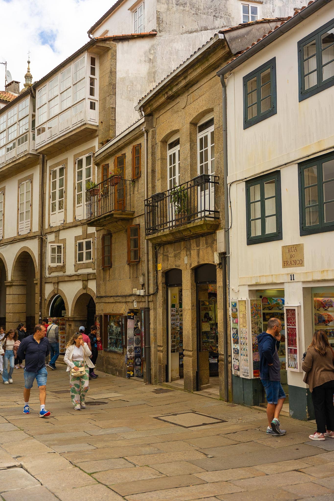 A granite lane in the old town of Santiago de Compostela with arcaded ground floors, glazed balconies and people walking on wet paving.