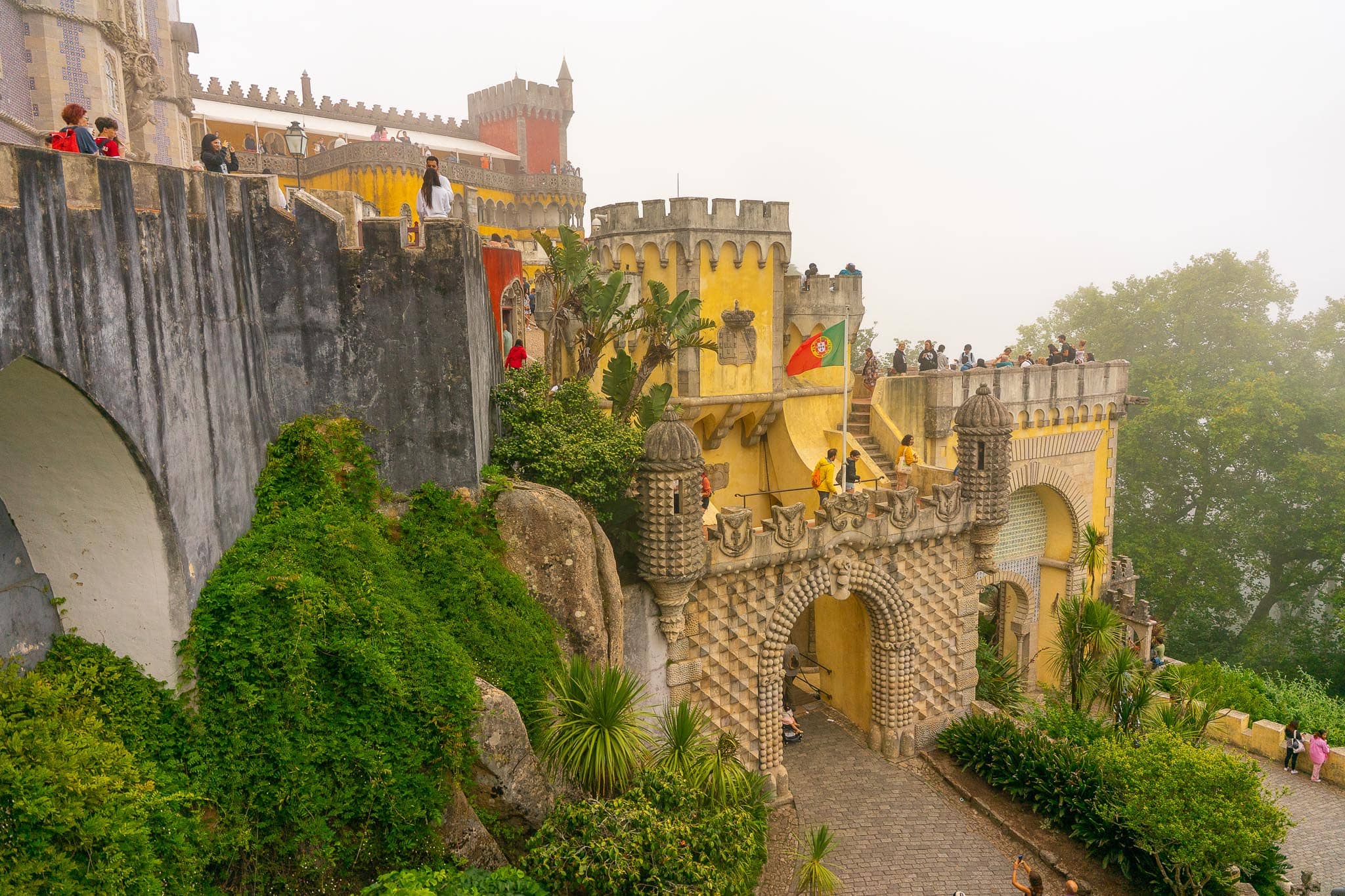 Stone ramparts and an arched gateway at the Pena Palace at Sintra, with dense green planting below and visitors on the walls above.