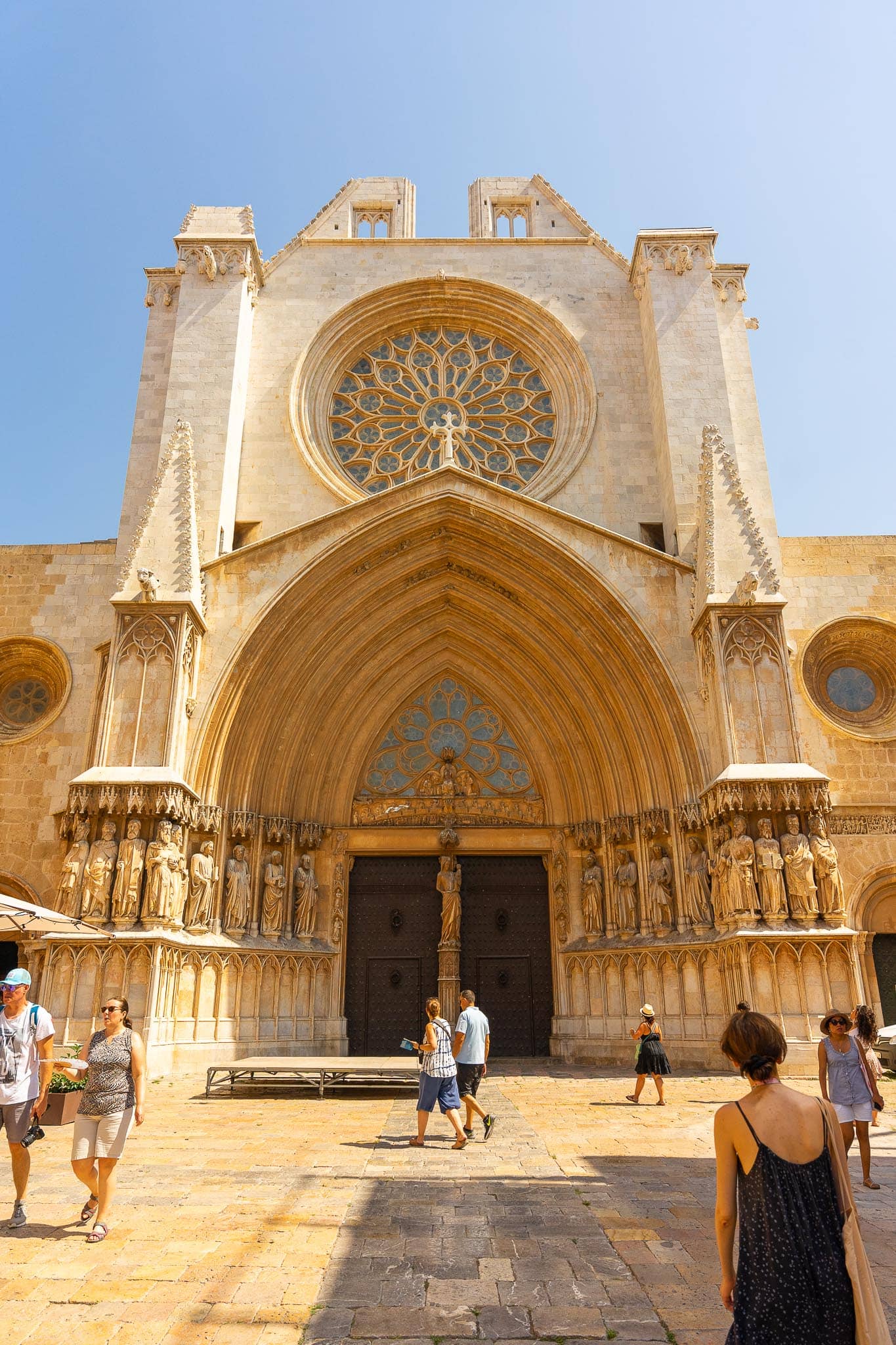 The unfinished stone facade of Tarragona cathedral with its large rose window, carved portal and flanking statues, people crossing the square below.