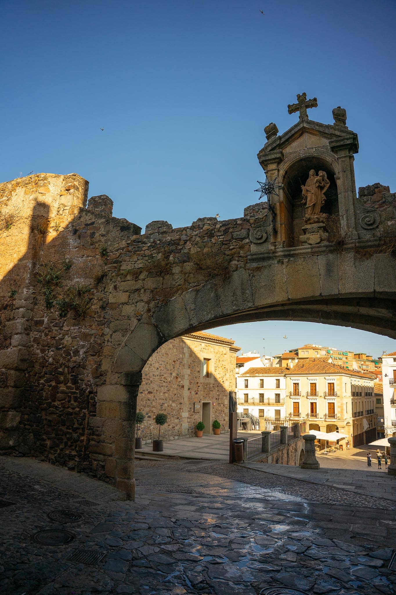 The Arco de la Estrella, a low stone arch breaching the walls of Caceres, a saint's statue in a niche above it, the modern town visible through the opening.