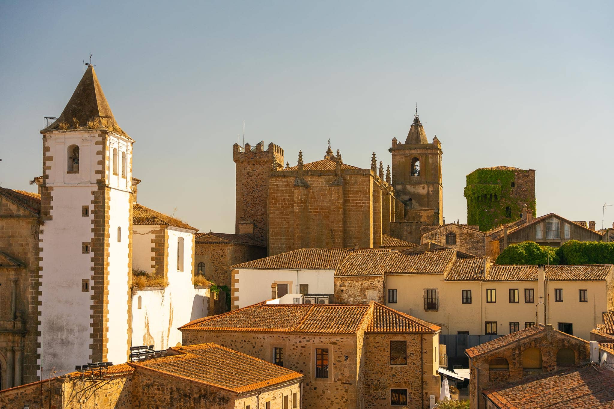 Terracotta rooftops, a whitewashed bell tower and the golden stone towers of the Ciudad Monumental in Caceres under late afternoon light.