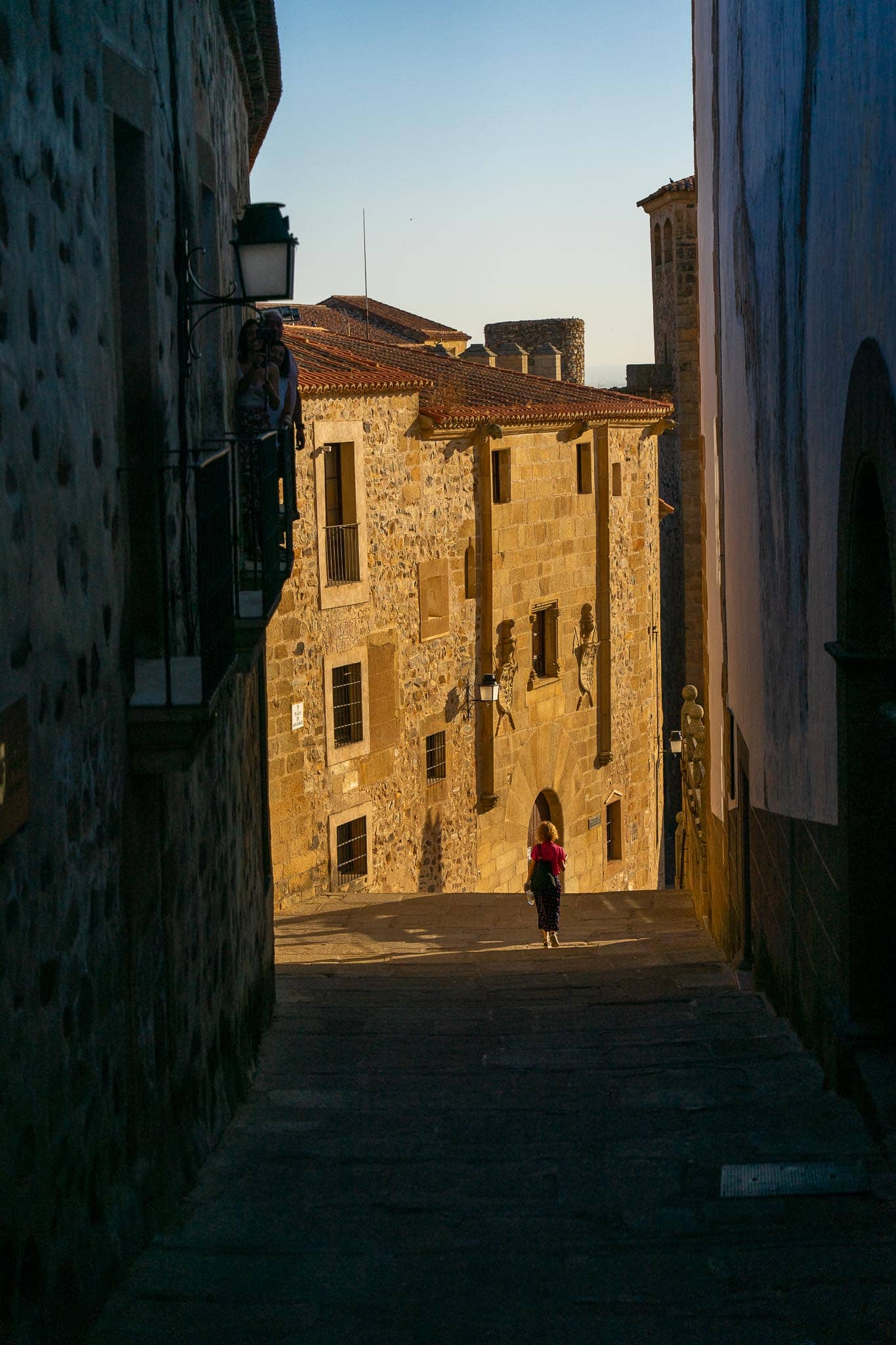 A narrow cobbled lane climbing between tall stone buildings in the old town of Caceres, a single figure in red walking up through deep shadow.
