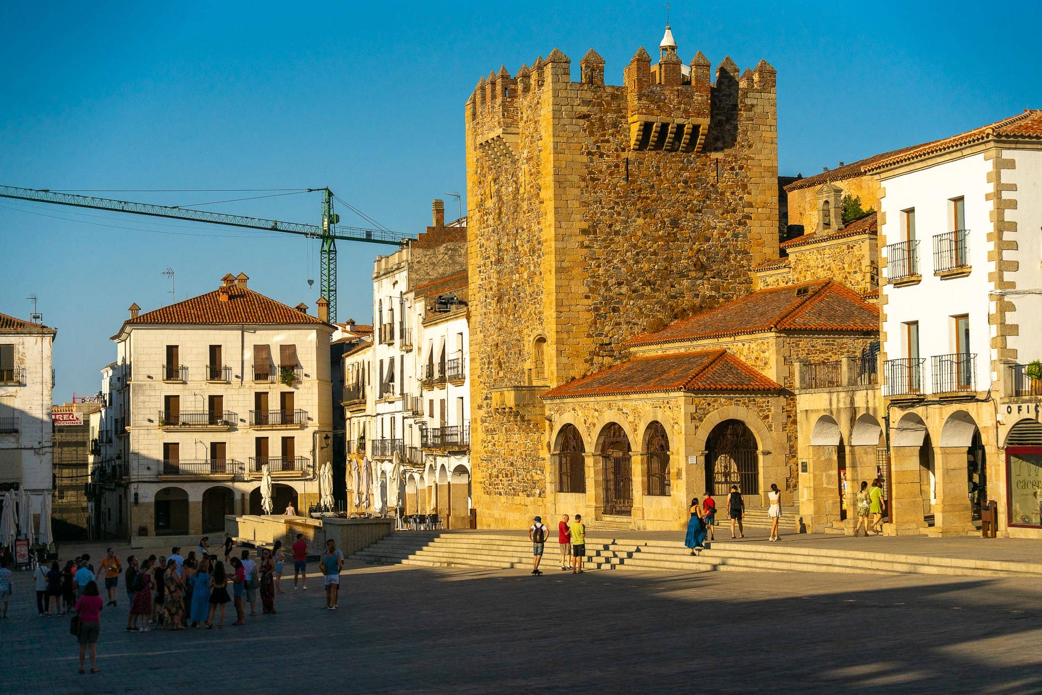 The battlemented Torre de Bujaco above the arcades of the Plaza Mayor in Caceres, lit gold by low evening sun, with people crossing the open square.