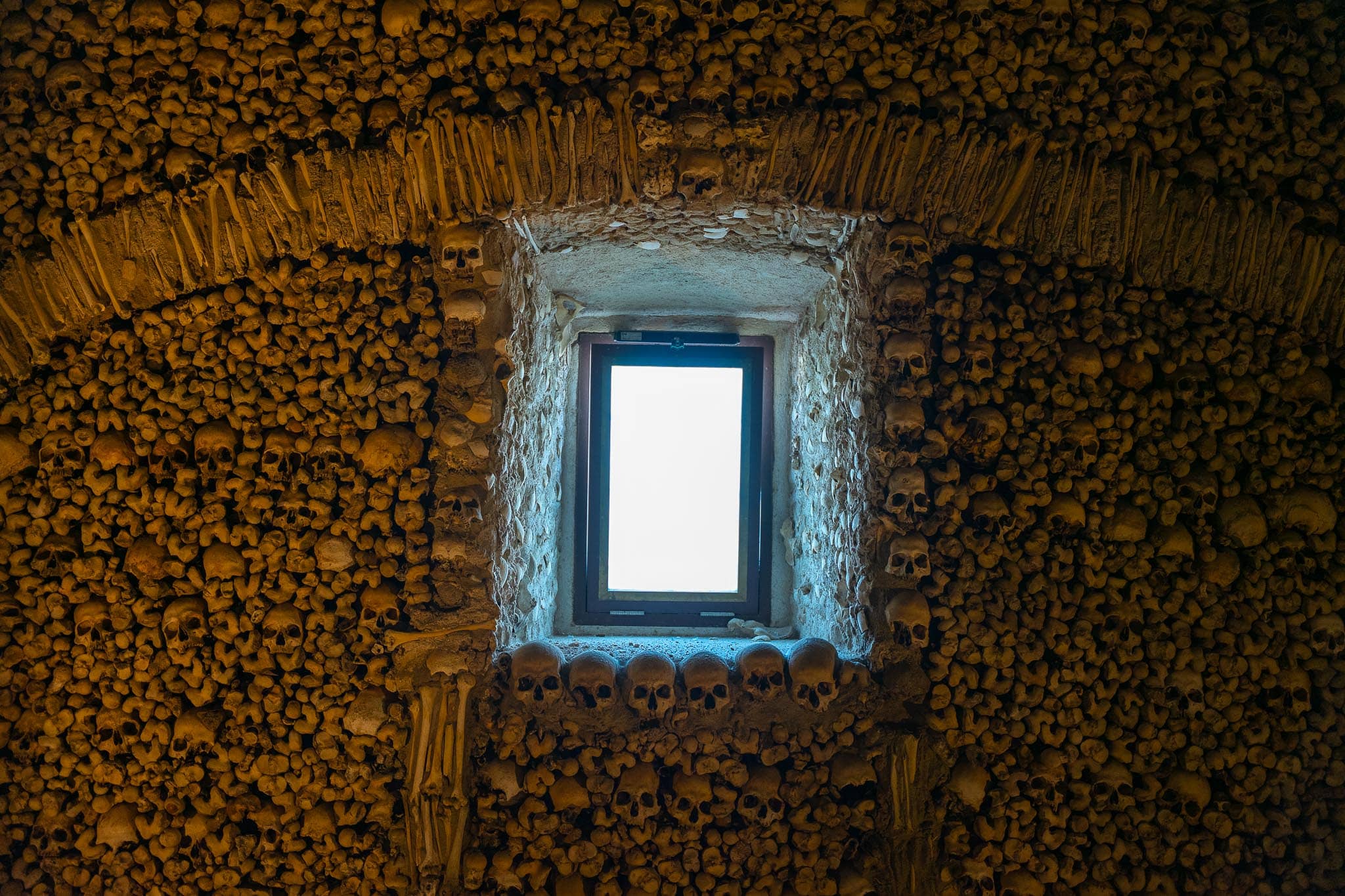 A window set into a wall faced entirely with human skulls and long bones in the Capela dos Ossos in Evora, lit by daylight from the opening.