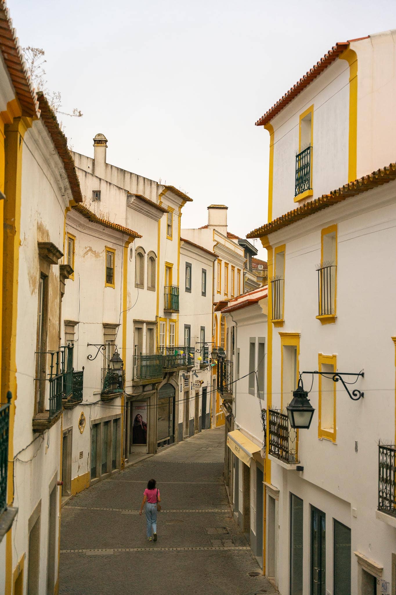 A narrow lane of white houses with yellow-painted borders and wrought-iron balconies in Evora, one figure walking away down the middle of the street.