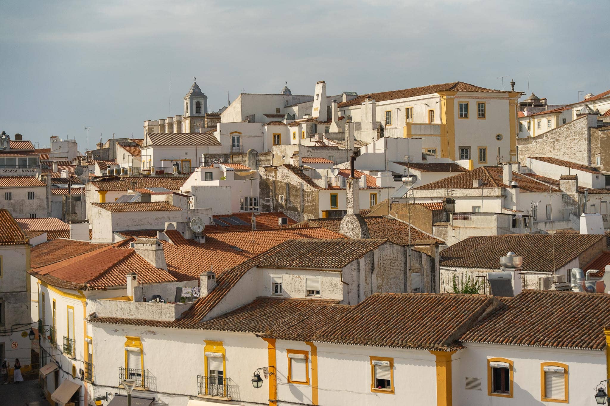 Whitewashed houses with terracotta roofs and yellow-trimmed windows packed across a slope in Evora under an overcast sky.
