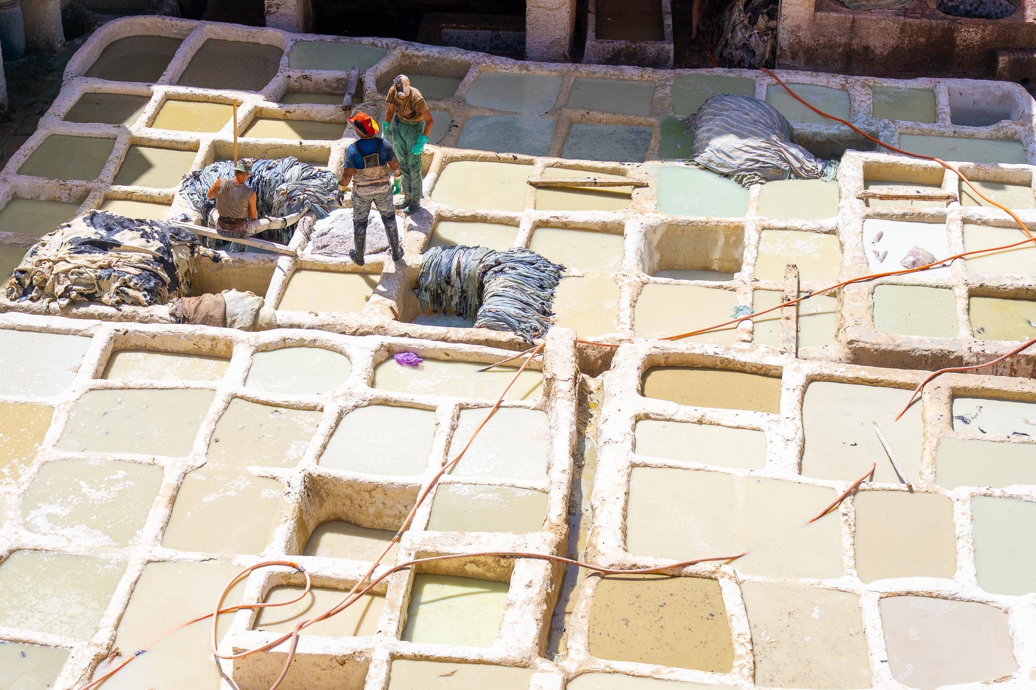 The Chouara tannery in Fez seen from above: a grid of white stone vats holding lime and pigeon-dung solutions, with hides stacked between them and two workers standing in the pits.