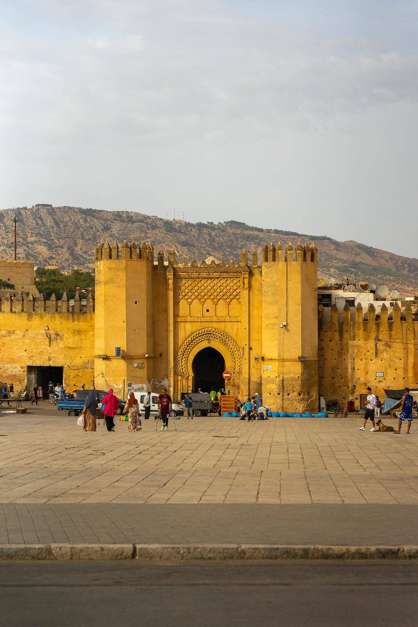 A monumental golden-stone gateway in the walls of Fez, three horseshoe arches under crenellations, with people crossing the open ground in front and dry hills behind.