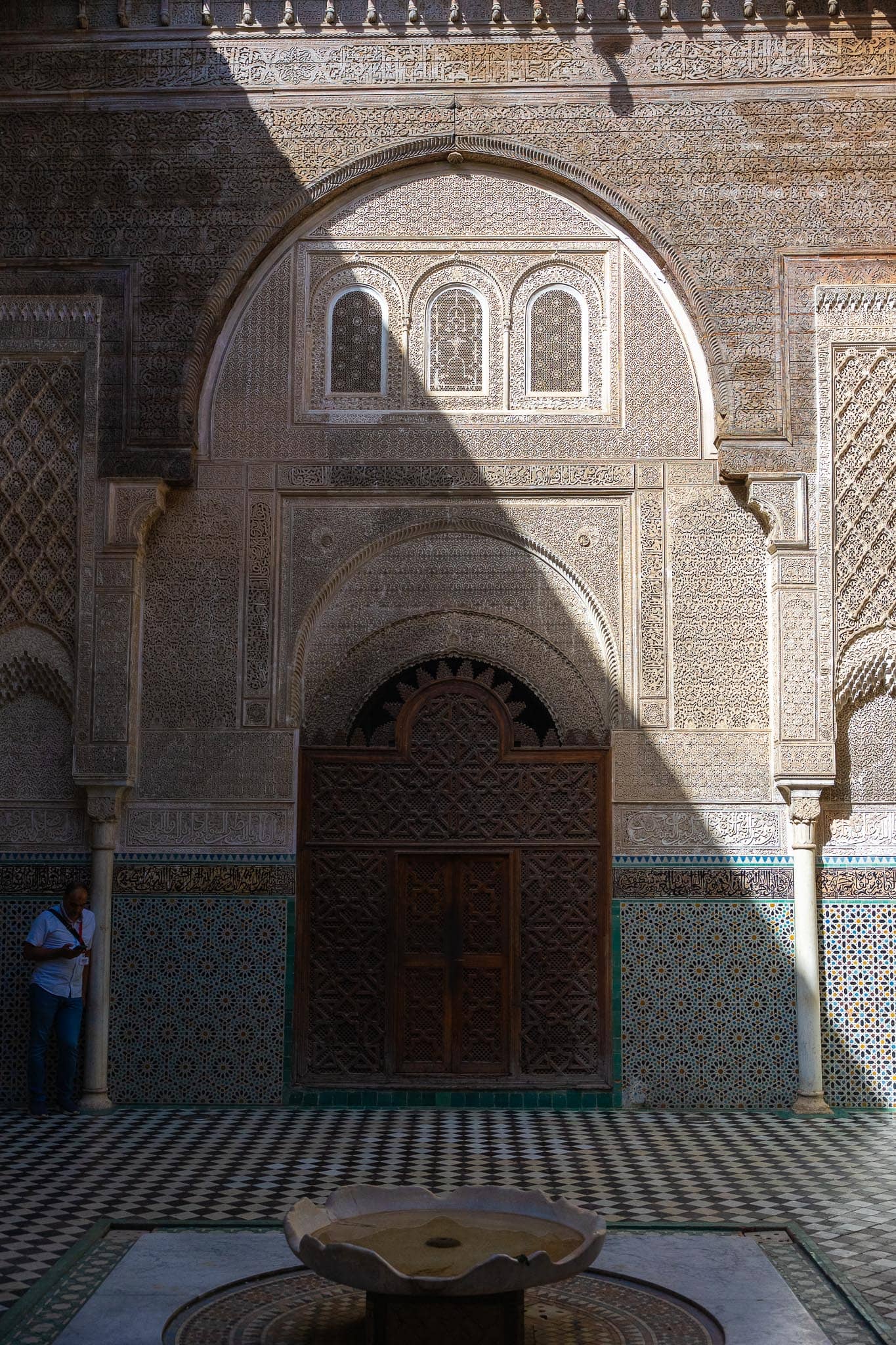 The courtyard of a medersa in Fez: a carved stucco horseshoe arch above a cedar door, zellij tilework below, and a low marble basin on the tiled floor.