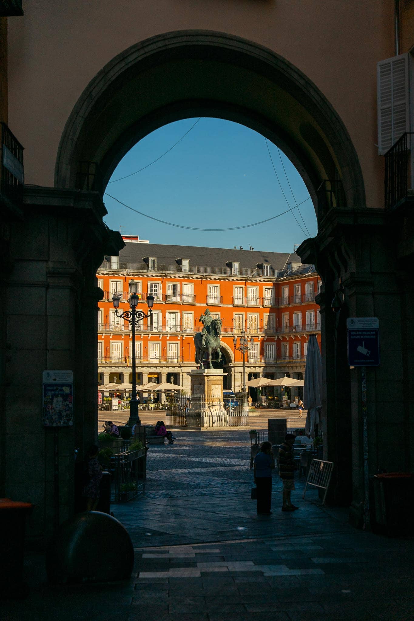 Looking through a dark stone archway into the Plaza Mayor in Madrid, its red-ochre facades and the bronze equestrian statue bright in the sun beyond.