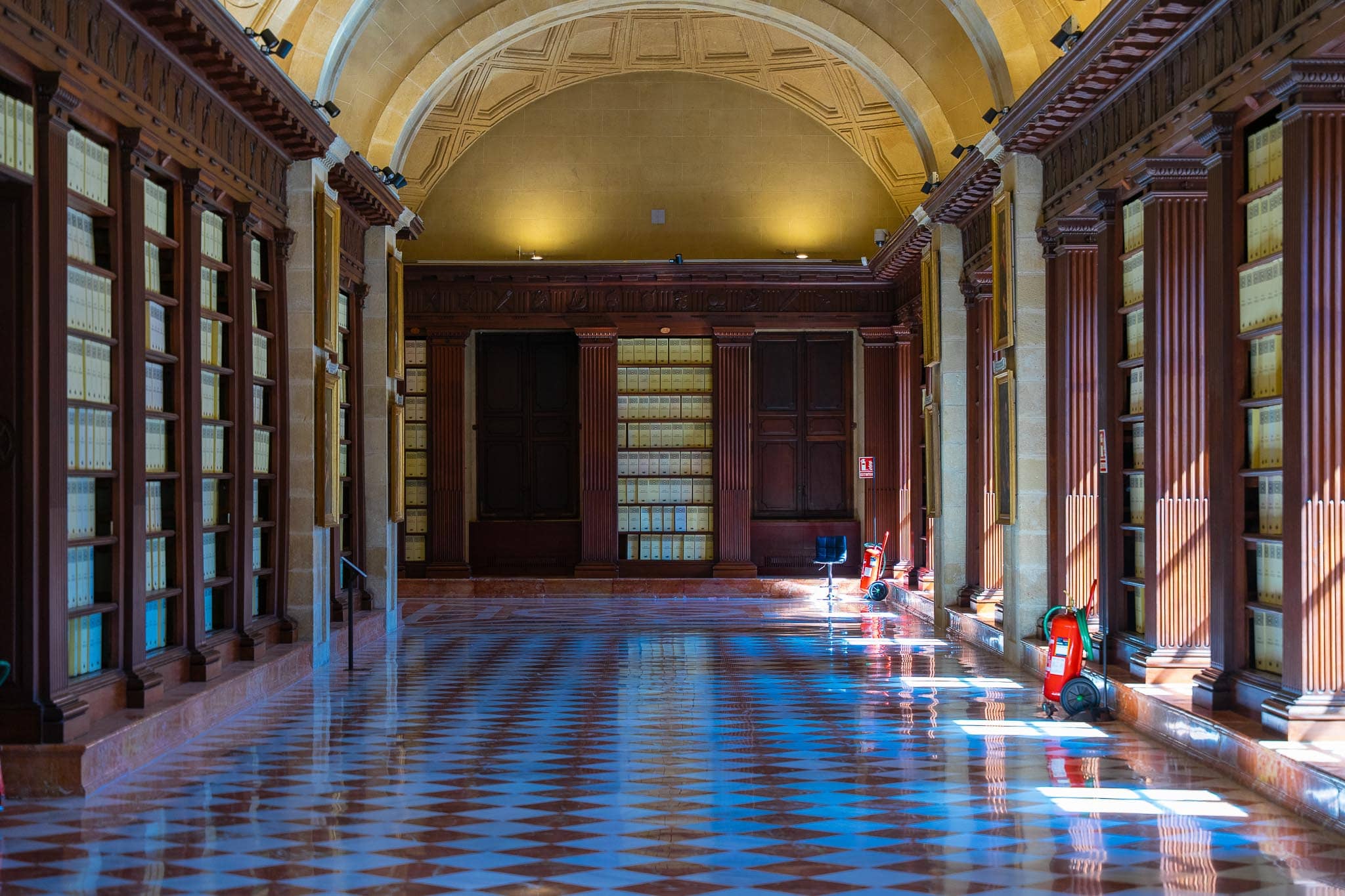 The vaulted upper gallery of the Archivo General de Indias in Seville, lined with uniform document boxes on mahogany shelving above a polished chequered marble floor cut by window light.
