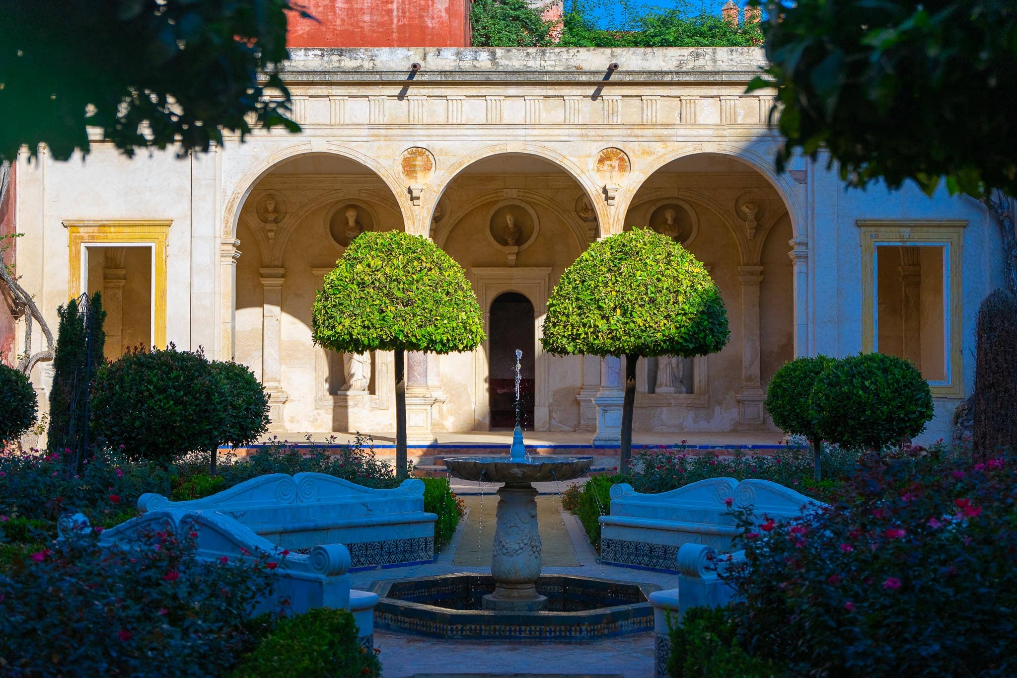 The garden loggia at Casa de Pilatos: three stone arches set with Roman busts in roundels, two clipped standard trees, an octagonal tiled fountain and marble benches backed with blue and white azulejos, among roses.