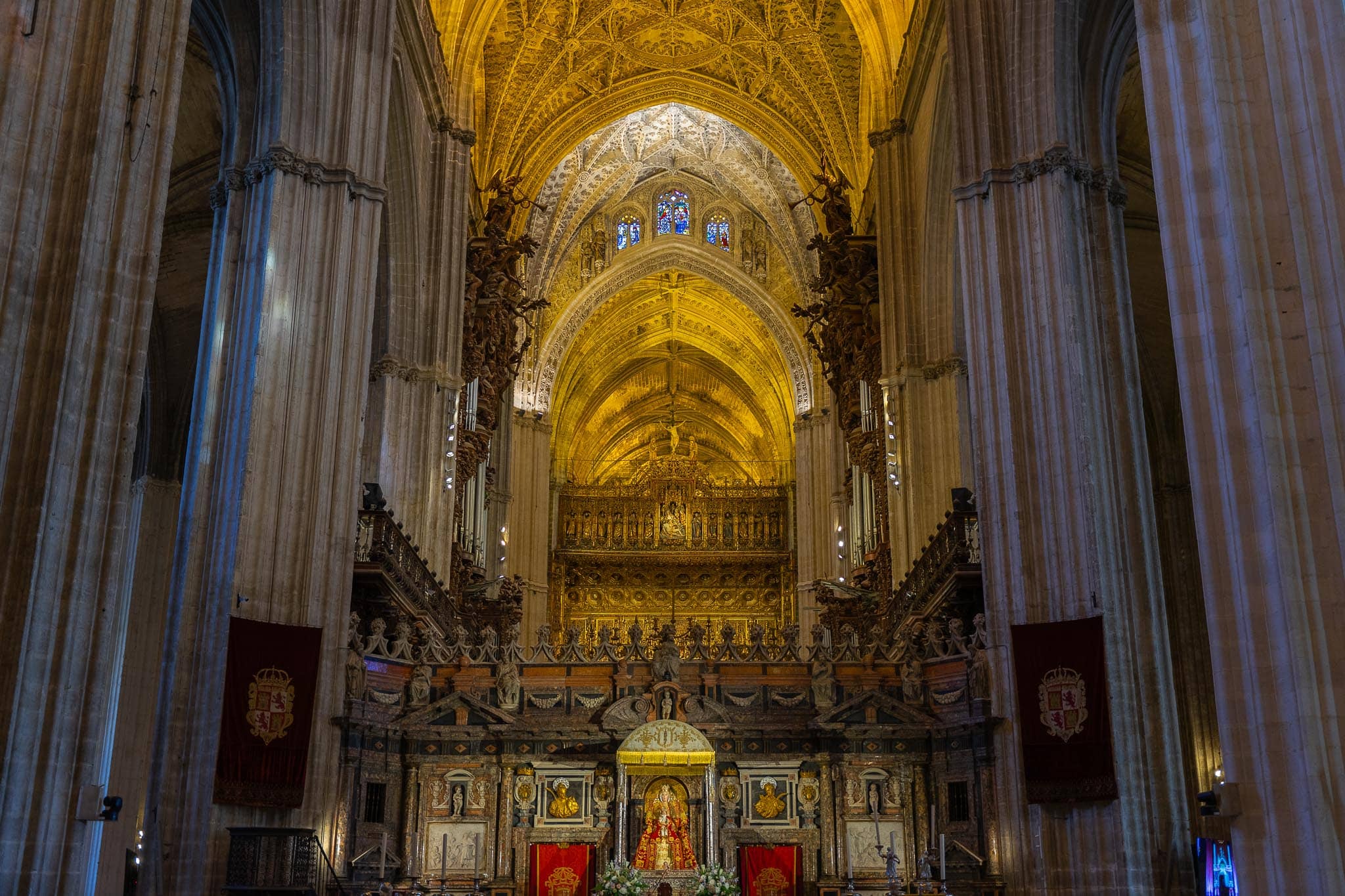 The interior of Seville Cathedral looking towards the Capilla Mayor, with its gilded altarpiece beneath tall Gothic vaults and two crimson heraldic banners hanging between the piers.