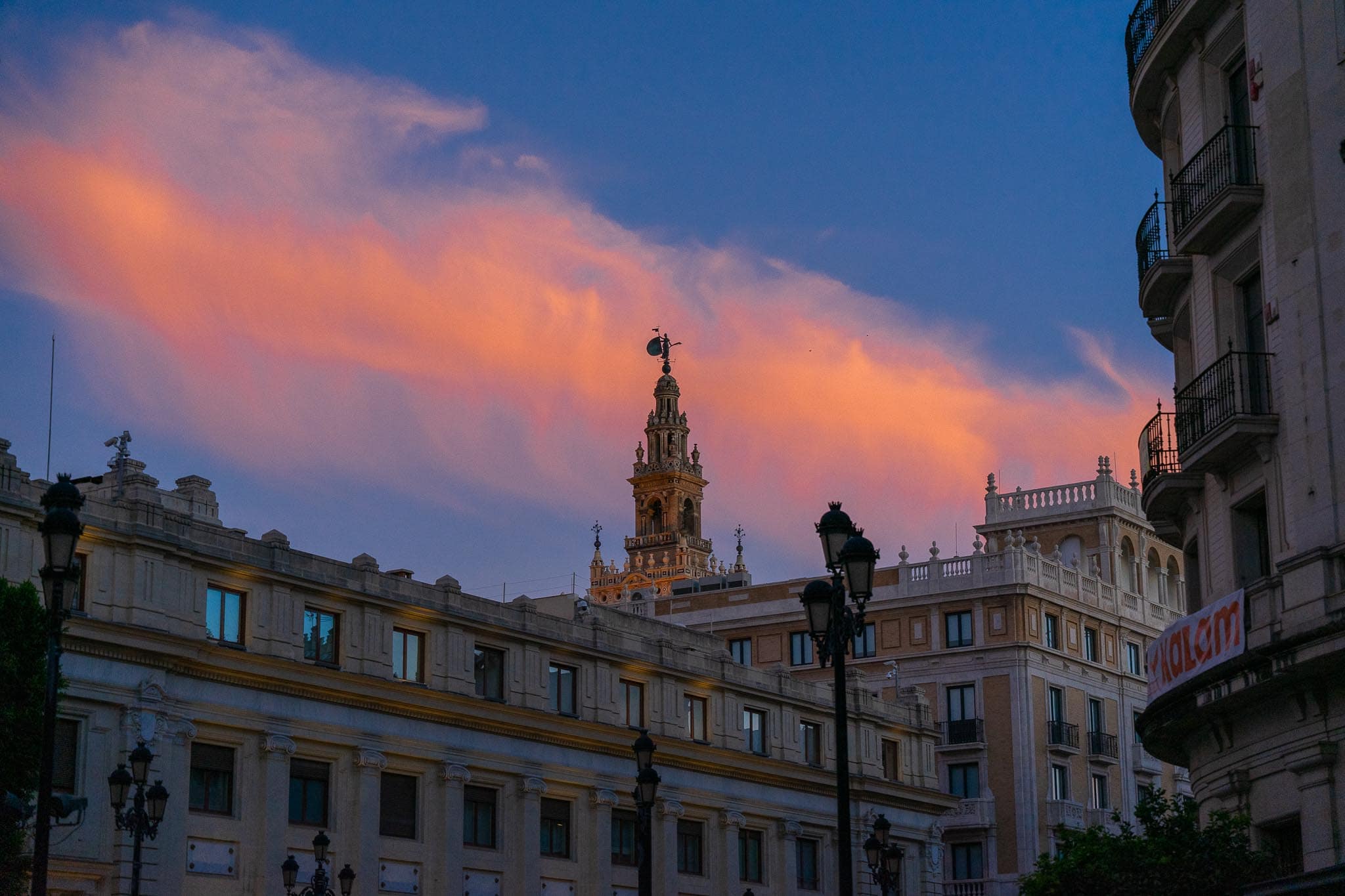 The Giralda bell tower catching the last of the light above a row of pale rooftops in Seville, beneath a bank of pink cloud.