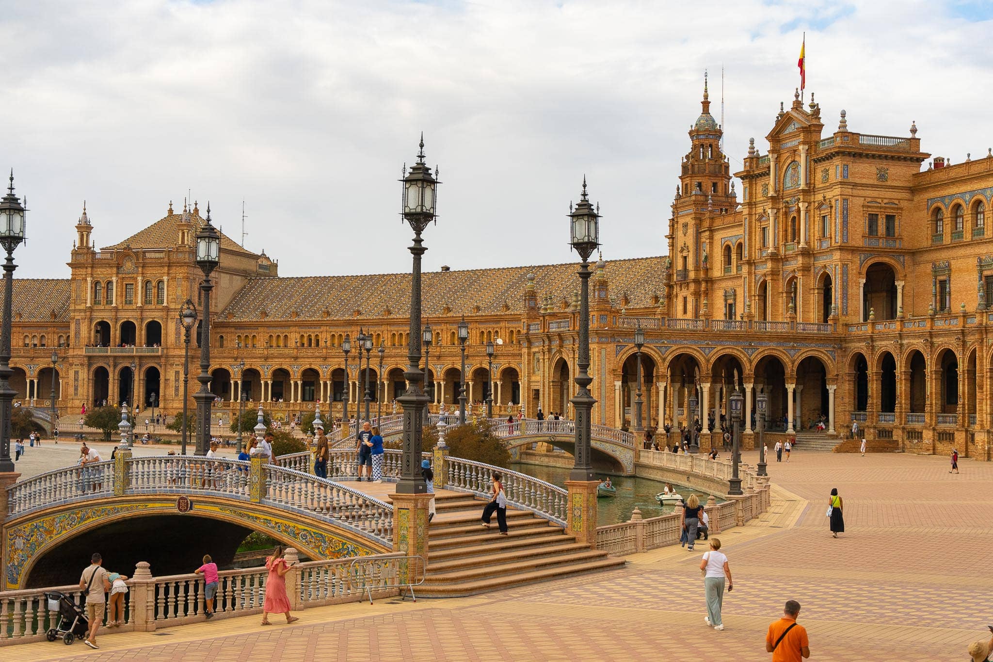 Plaza de España in Seville: tiled balustraded bridges crossing the canal in front of the curved brick arcade and its tiled tower, with a scattering of visitors on the paving.