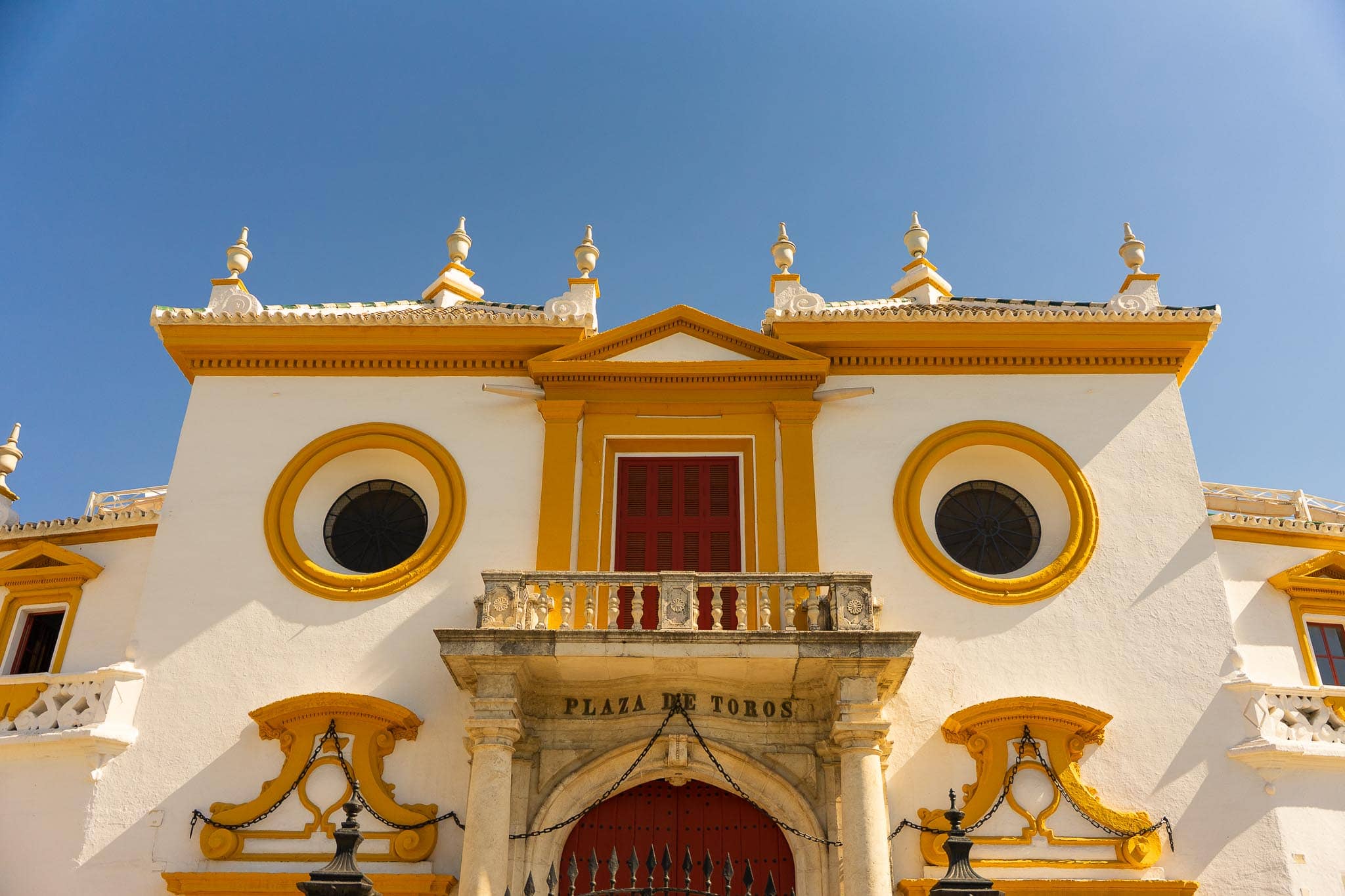 The principal façade of the Plaza de Toros de la Real Maestranza in Seville: white walls with ochre-yellow mouldings, two circular windows, a red shuttered balcony door, and PLAZA DE TOROS carved into the stone lintel.