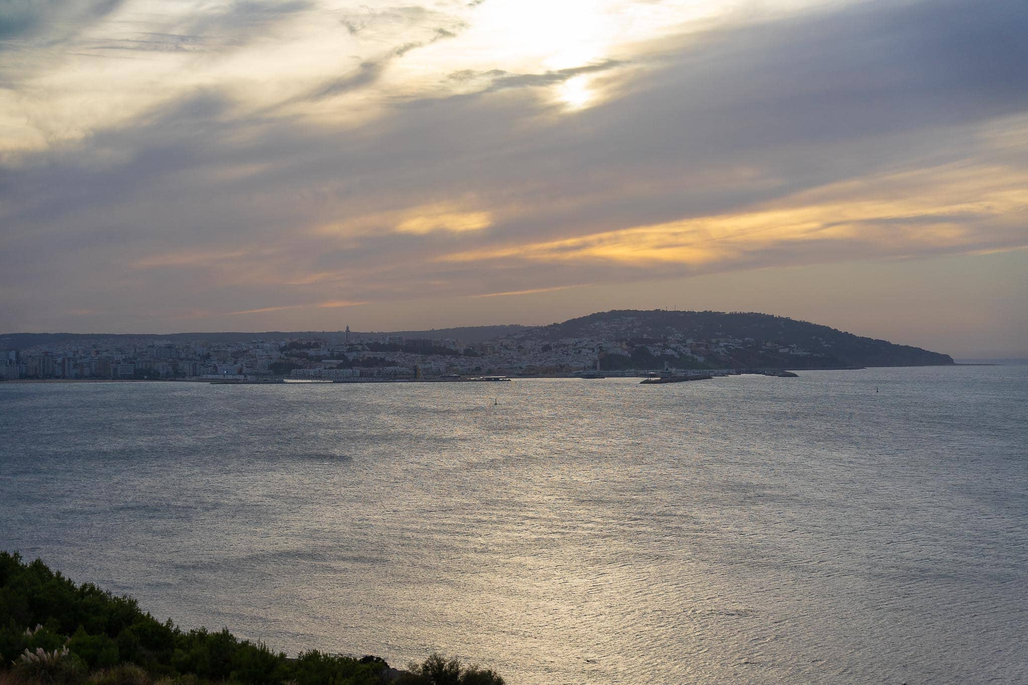 The Bay of Tangier at sunset: flat silver water, a long low headland running out to sea, and broken cloud with the sun behind it.