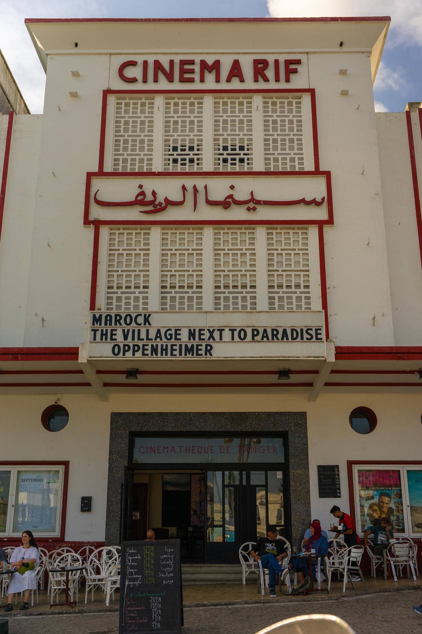 The Cinema Rif on the Grand Socco in Tangier: a cream art-deco frontage with red lettering in Latin and Arabic script, film titles on the marquee and cafe tables outside.
