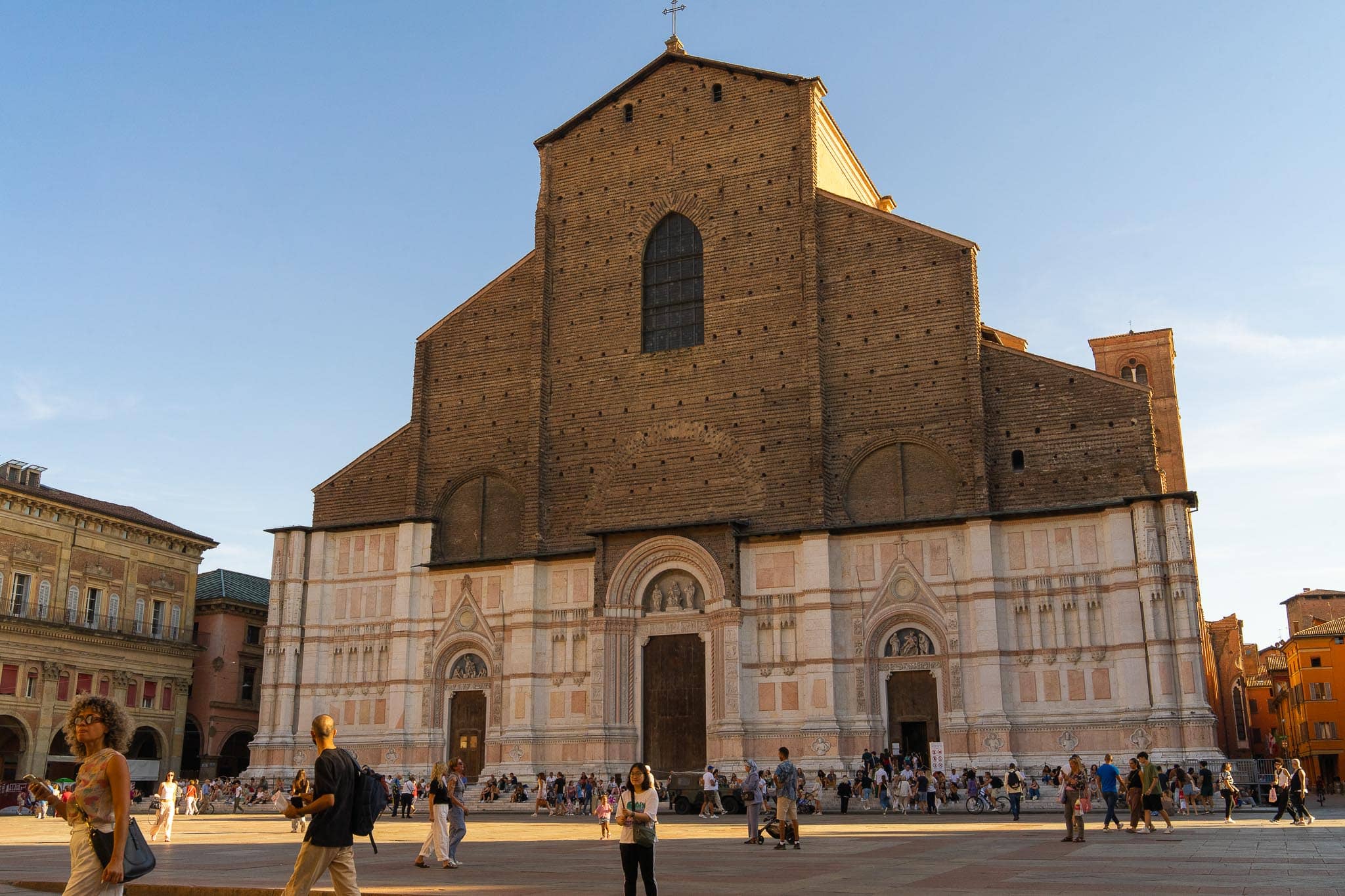 The west front of the Basilica di San Petronio in Bologna: marble facing across the lower half, bare brick above where the cladding was never finished, with people crossing the square.