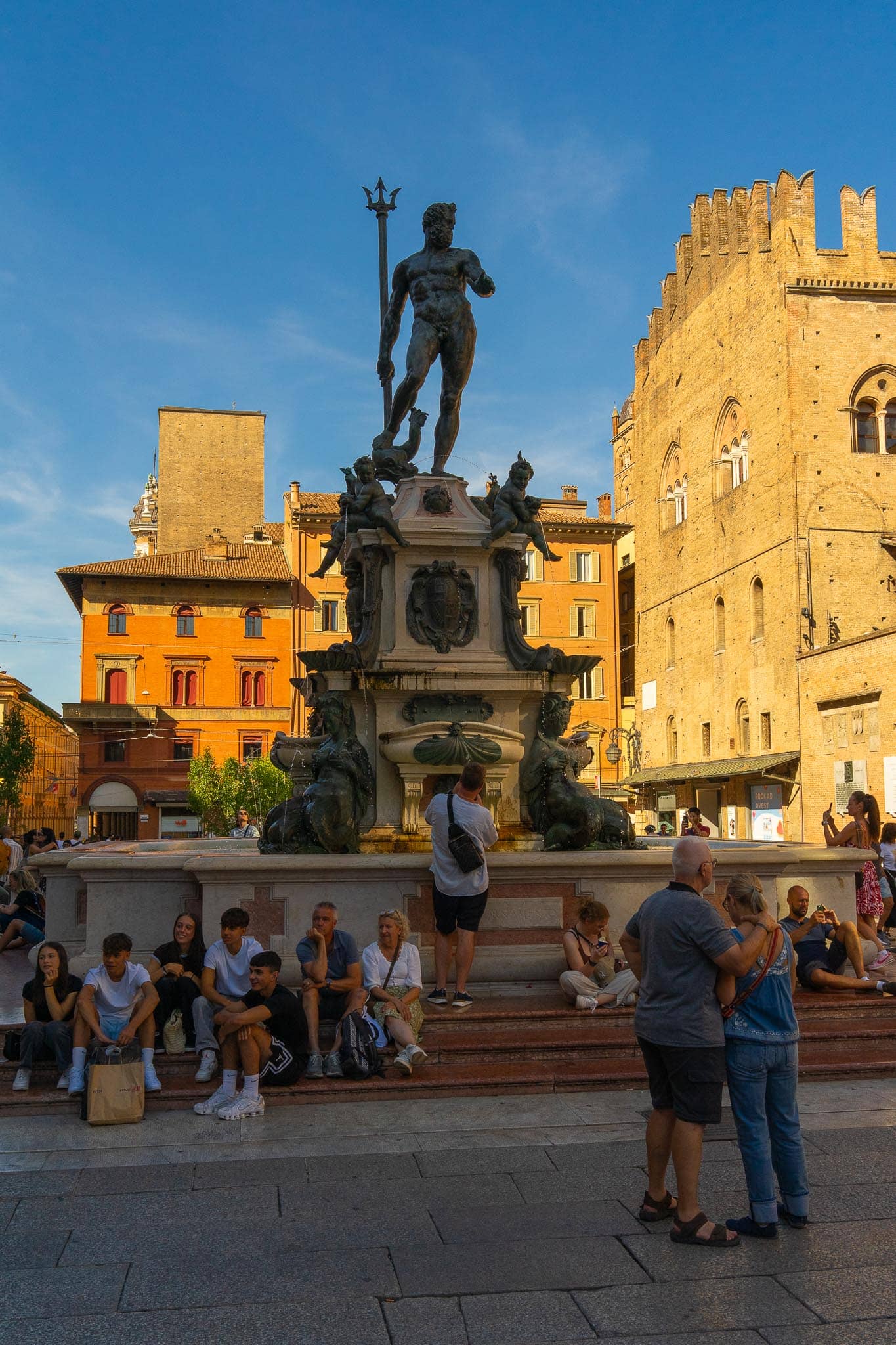 The bronze Fontana del Nettuno in Bologna, Neptune with his trident above the basin, backed by the brick and ochre palazzi of Piazza Maggiore, with people sitting on the fountain steps.