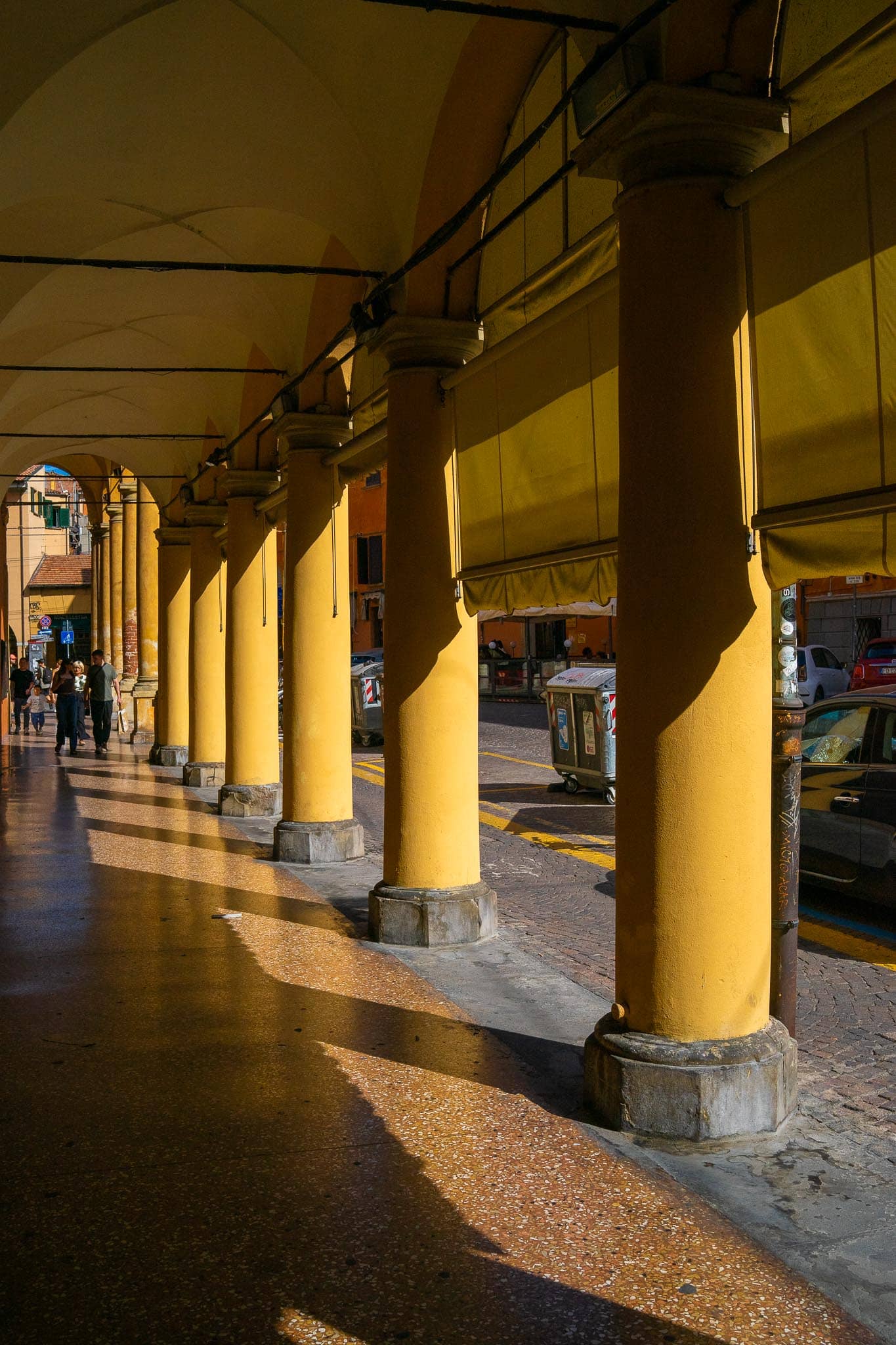 A Bologna portico in low evening sun: a row of ochre-painted columns on stone bases carrying the vaulted bays overhead, long shadows thrown across the terrazzo pavement, parked cars at the kerb beyond, and a few people walking far down the arcade.
