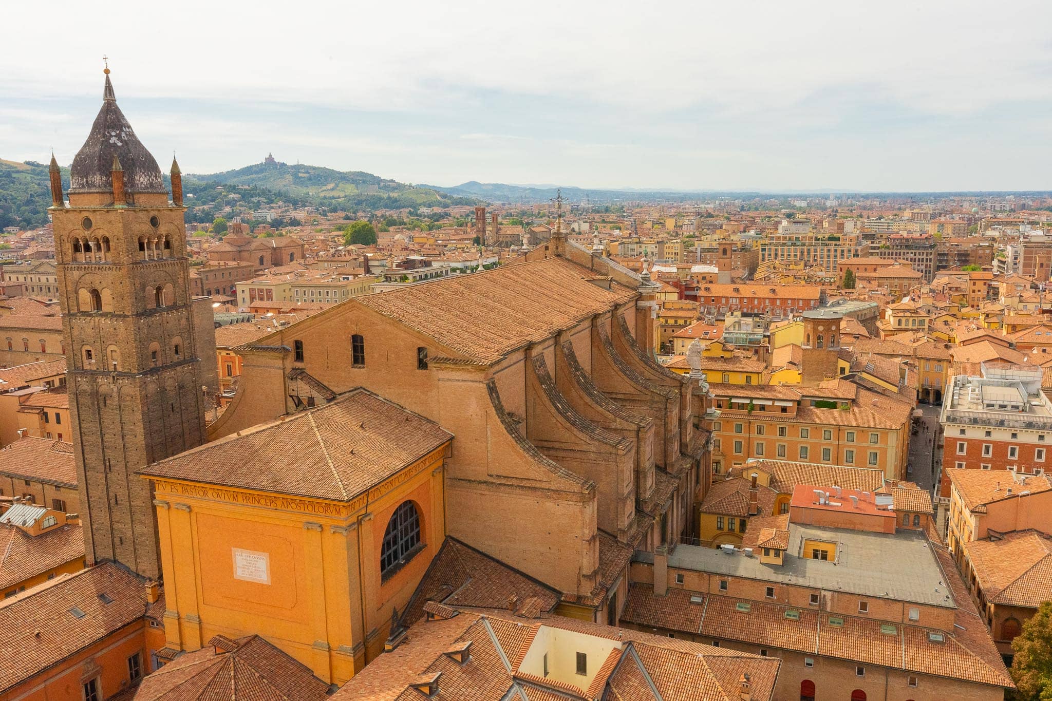 Bologna from above: a sea of terracotta roofs, a tall brick campanile in the middle distance, and the wooded hills with the Sanctuary of San Luca on the skyline.