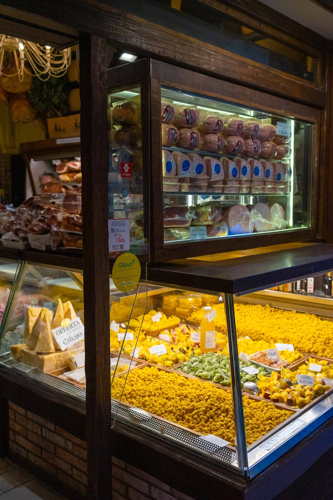 A lit delicatessen window in Bologna stacked with cured meats and cheeses above trays of fresh yellow egg pasta and filled tortellini.