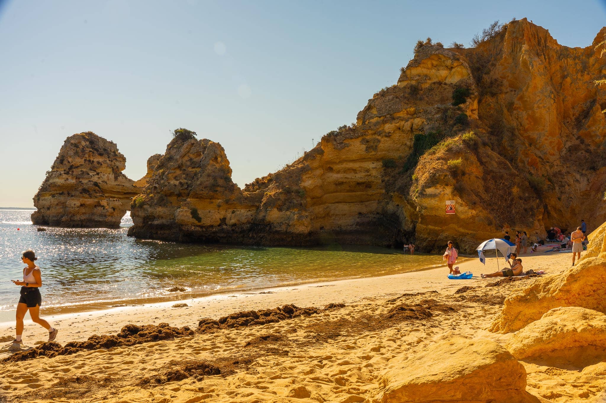 A small sandy beach enclosed by golden cliffs and a pierced rock stack near Lagos, with a few people and a parasol on the sand in early light.