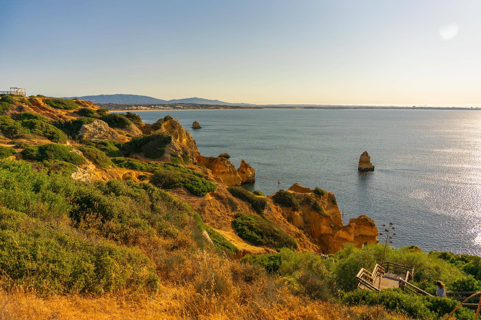 A scrub-covered headland of golden cliffs dropping into a calm sea near Lagos, with a lone rock stack offshore and the coast fading into haze.