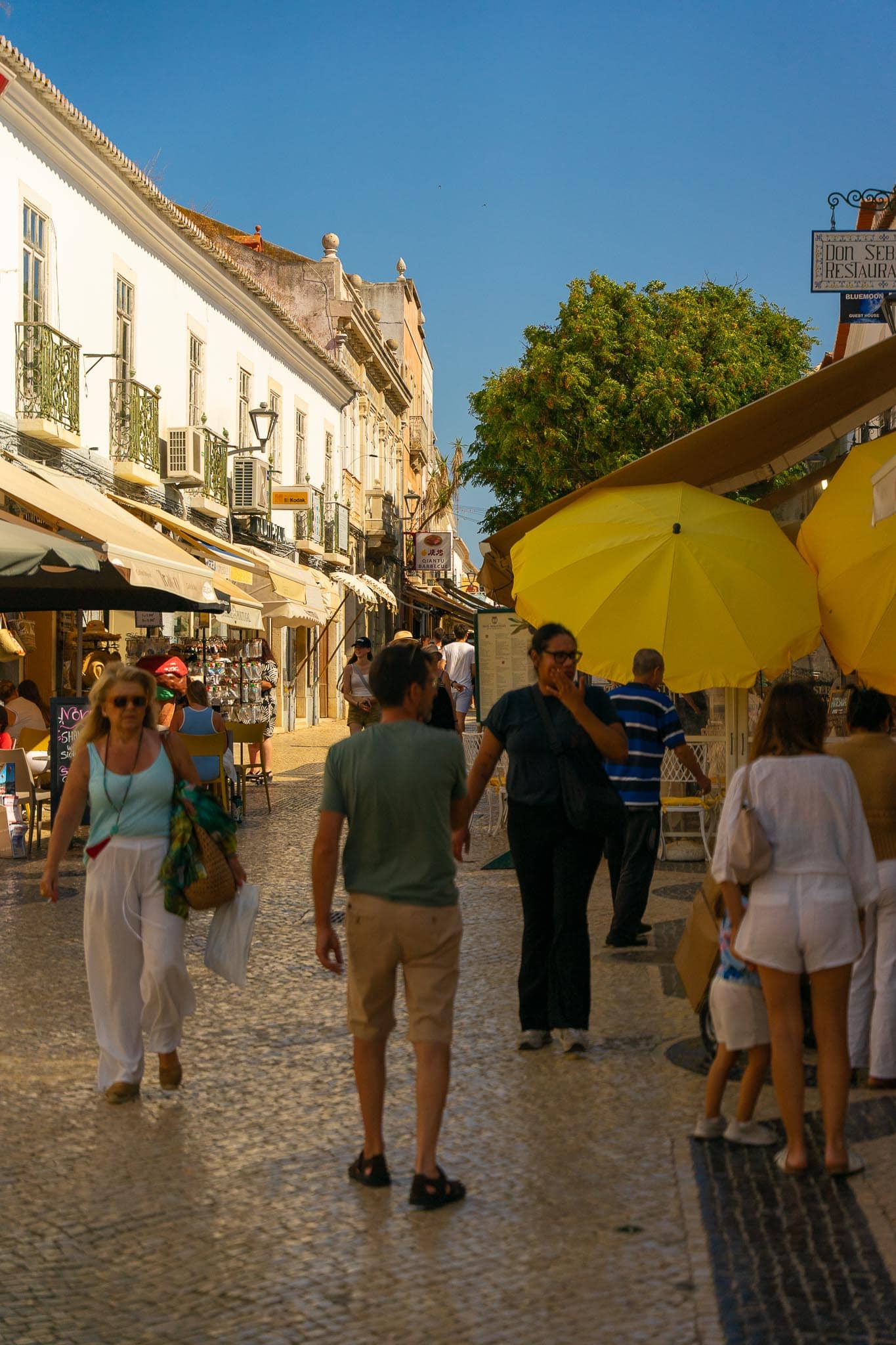 A pedestrian street in the old town of Lagos with whitewashed buildings, yellow cafe parasols and people walking on patterned calcada paving.