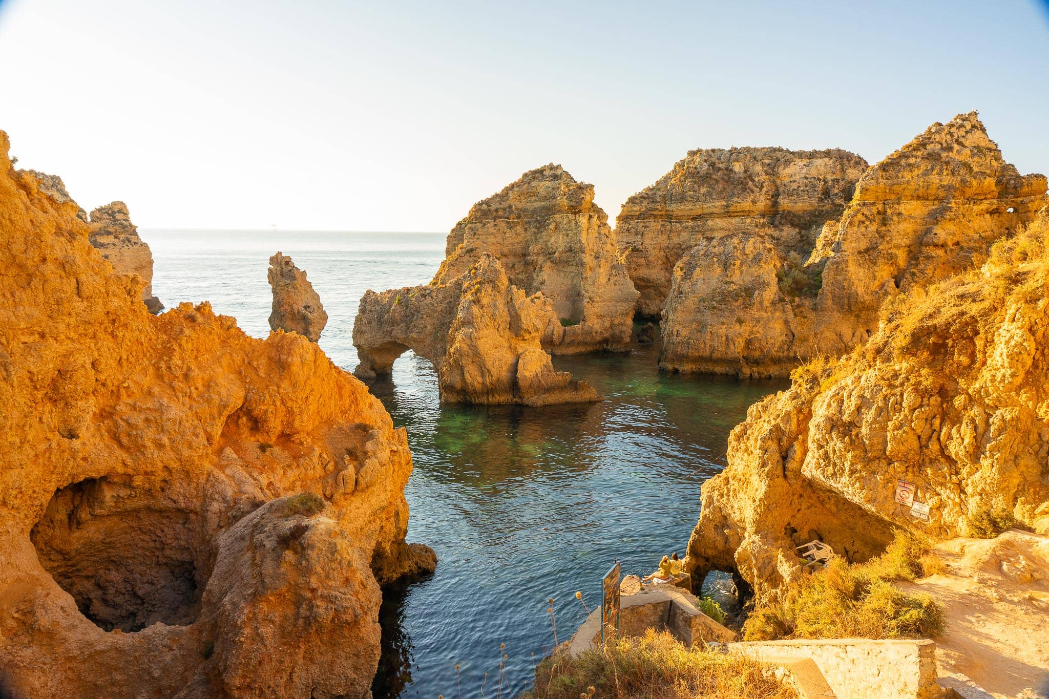 Golden sandstone sea stacks and natural arches at Ponta da Piedade near Lagos, with clear turquoise water between them and a stepped path at the right.