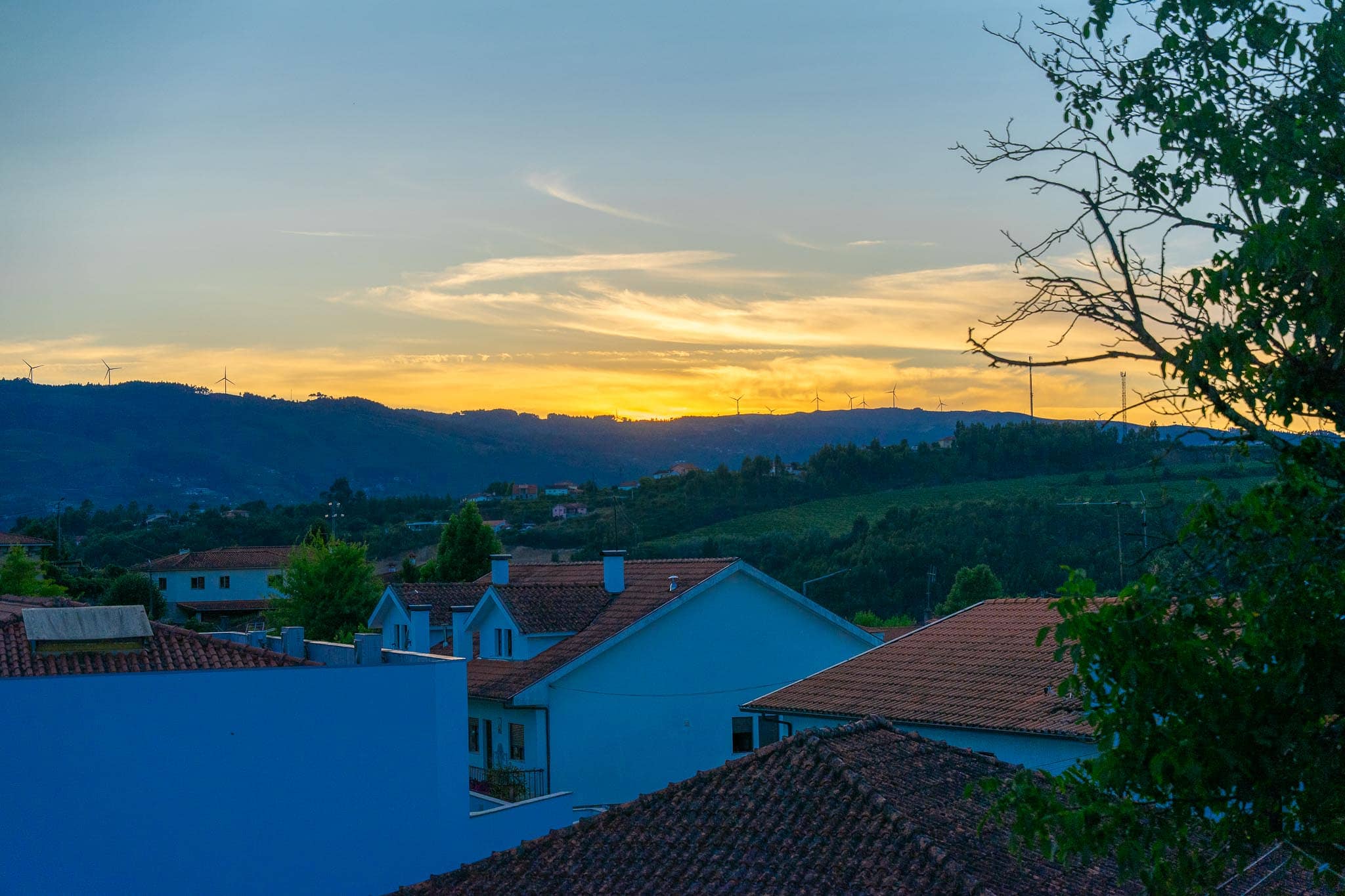 Sunset over wooded green hills above the tiled rooftops of houses in Mondim de Basto, with bands of yellow light along the ridgeline.