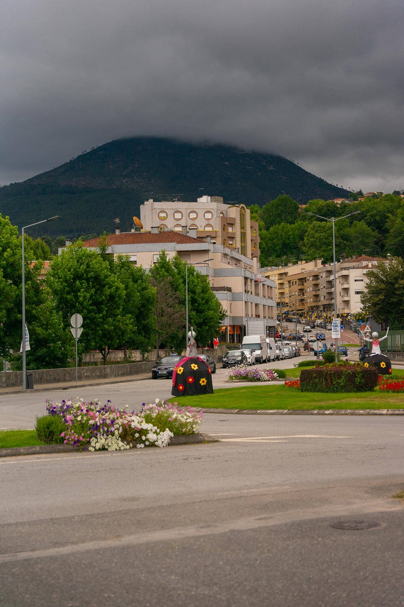 A conical wooded mountain under heavy cloud rising above the low buildings, trees and roadside flowerbeds of Mondim de Basto.