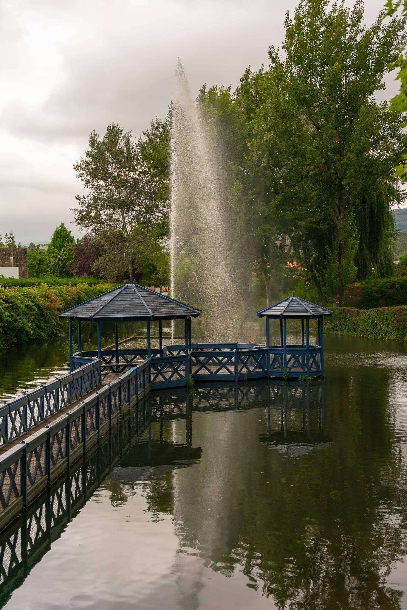 Two roofed pavilions on a railed walkway over a still green lake in Mondim de Basto, with a tall fountain jet and poplars behind.