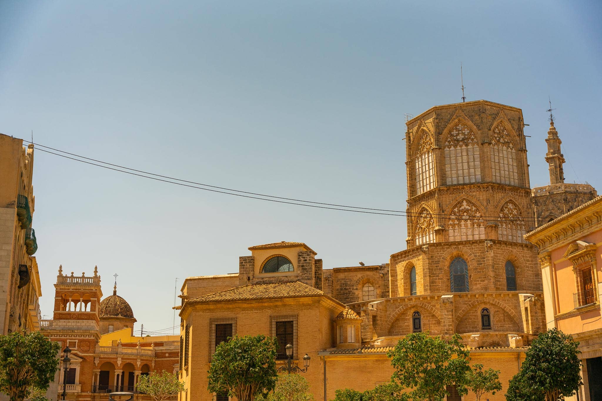 The octagonal lantern tower and domes of Valencia cathedral above ochre rooftops and orange trees in warm low evening light.