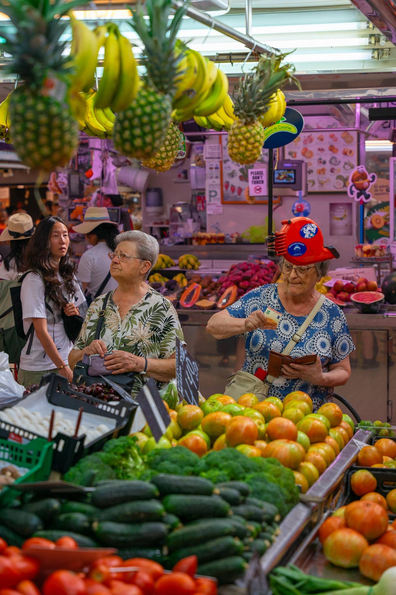 A fruit and vegetable stall in Valencia's Mercado Central, tomatoes and citrus banked up under hanging pineapples and bananas, with shoppers at the counter.