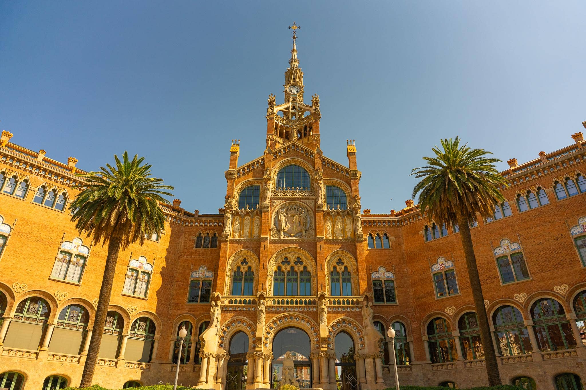 A pavilion of the Hospital de Sant Pau in Barcelona: red brick and stone modernista facade with mosaic panels, sculpture and a spire, flanked by two tall palms.