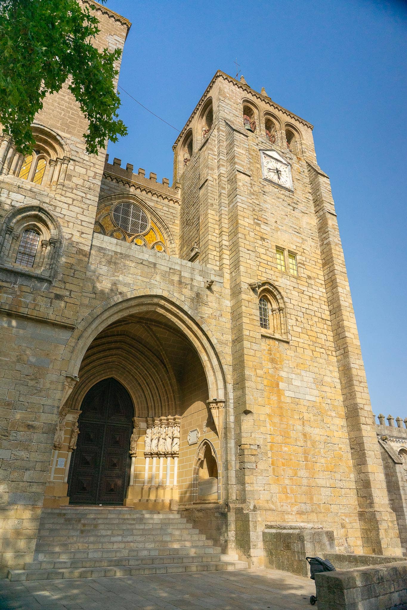 The granite west portal and clock tower of Evora cathedral, a deep Gothic arch flanked by carved apostles, with battlemented parapets above.