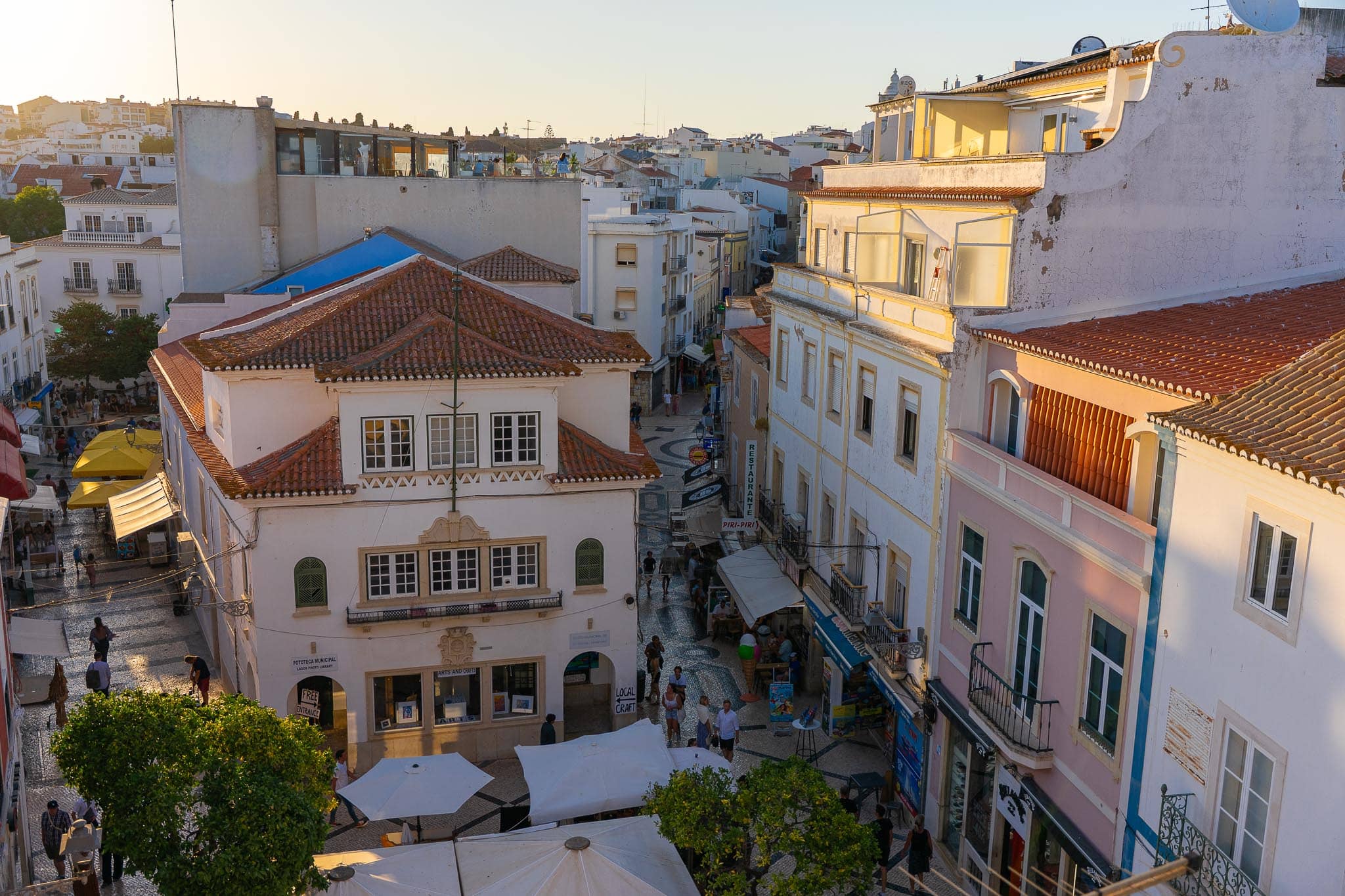 The old town of Lagos from above: white and pastel buildings with terracotta roofs, market awnings in the street below and the town running down to the sea.