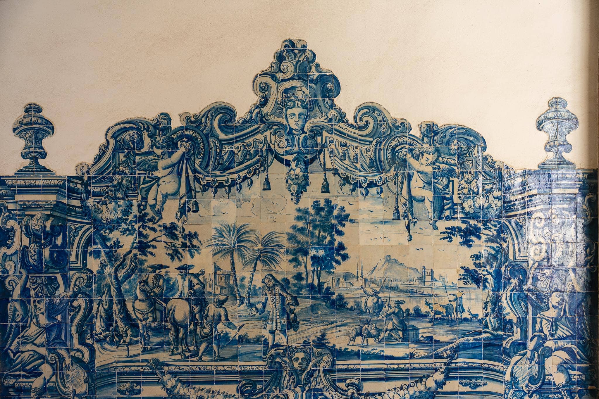A large blue-and-white azulejo panel in Lisbon showing a scrolled baroque cartouche above a landscape with figures, palm trees and a horseman.