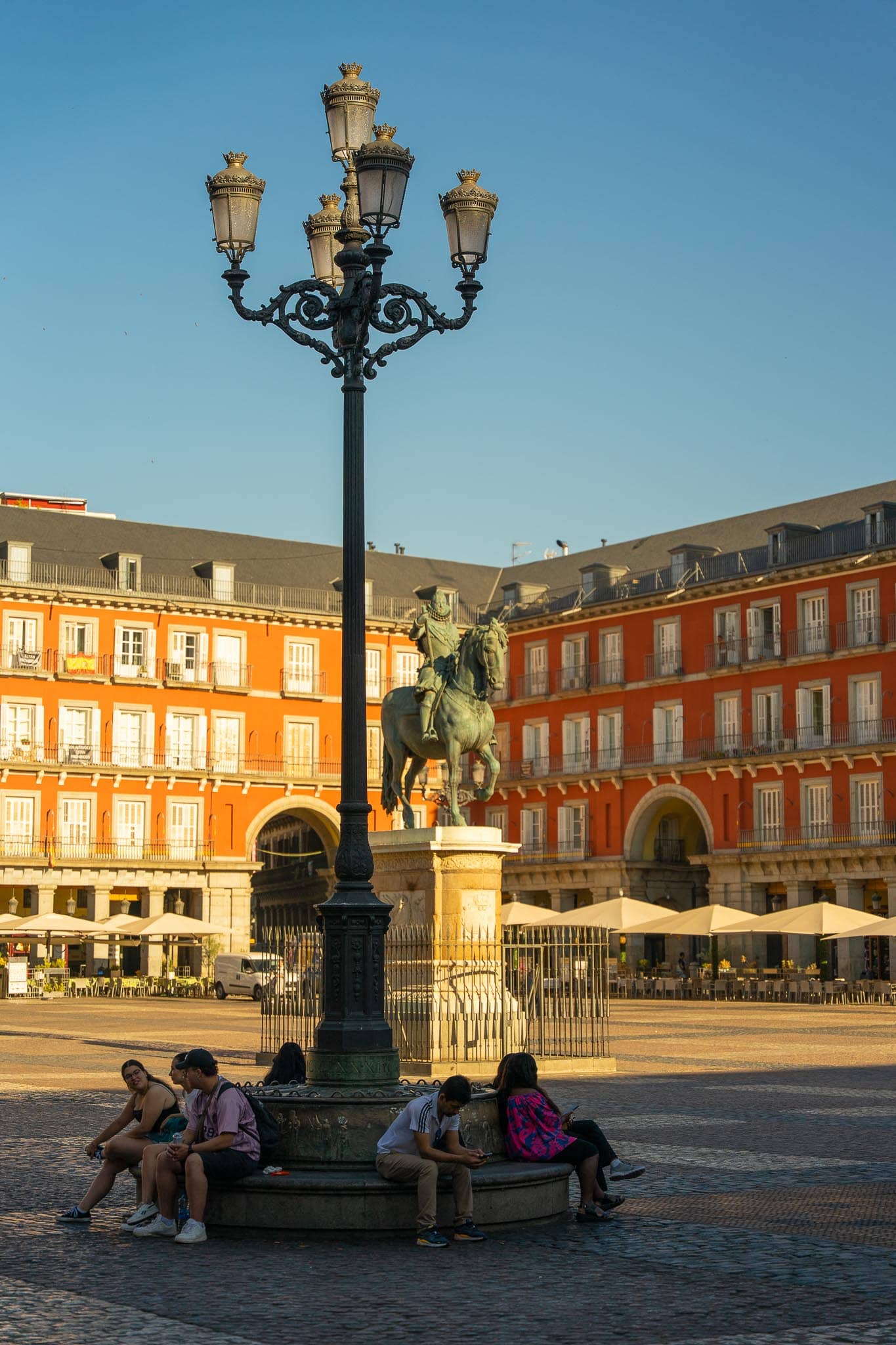 The Plaza Mayor in Madrid in low sun: red-ochre arcaded facades, an ornate iron lamp standard and the bronze equestrian statue, with people sitting at its base.