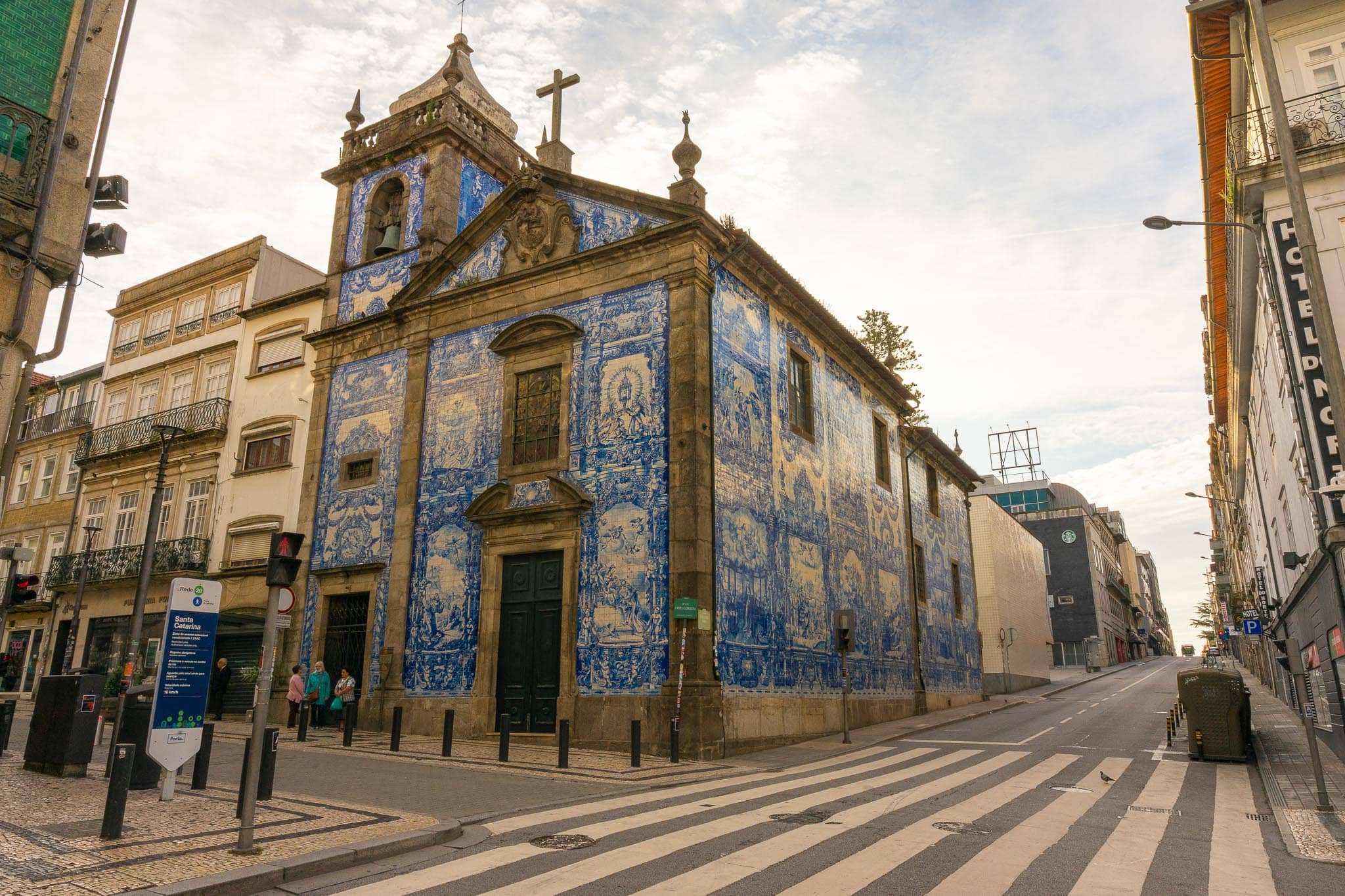The Capela das Almas in Porto, its exterior walls faced entirely in blue-and-white azulejo panels depicting saints' lives, on a street corner with tram rails and a crossing.