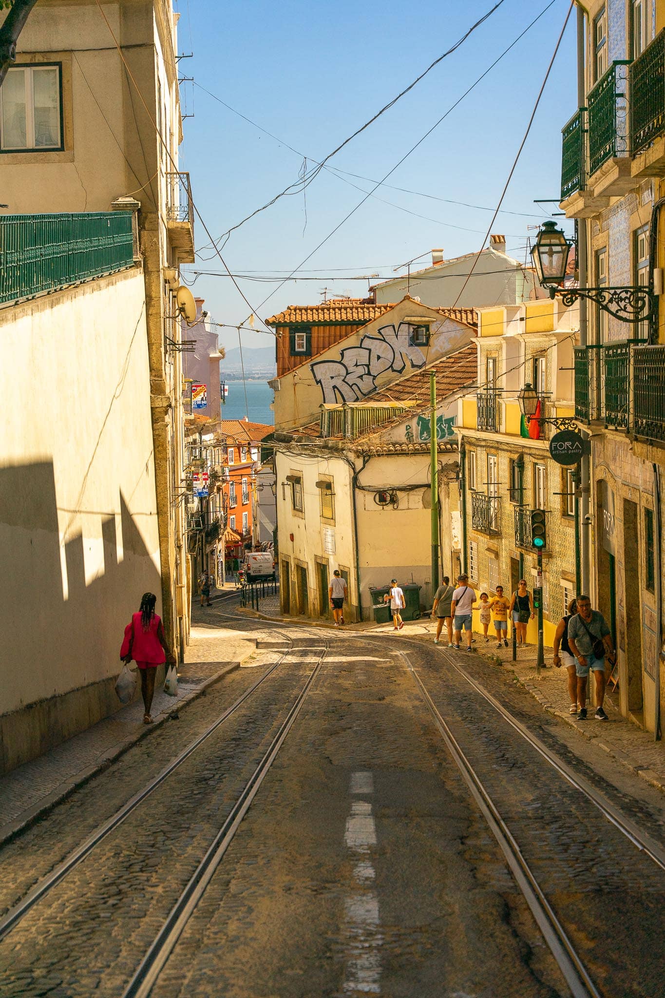 A narrow Lisbon lane with tram rails set into the paving, washing lines and overhead wires, dropping downhill towards the river.