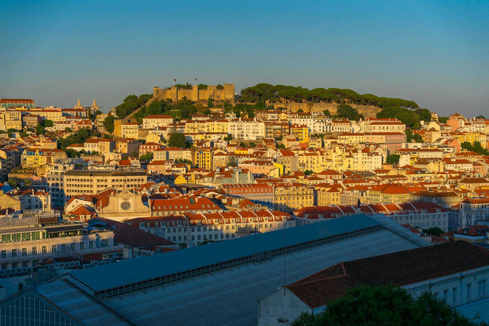Terracotta rooftops of Alfama in Lisbon packed up the hillside towards the castle walls, lit warm by low sun.