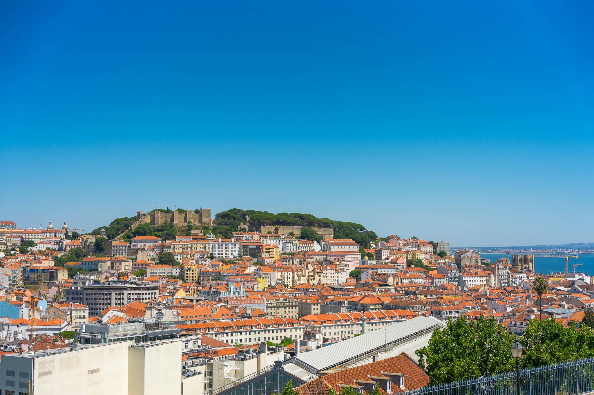 The walls of the Castelo de Sao Jorge on its wooded hill above the terracotta rooftops of central Lisbon, with the Tagus estuary beyond.