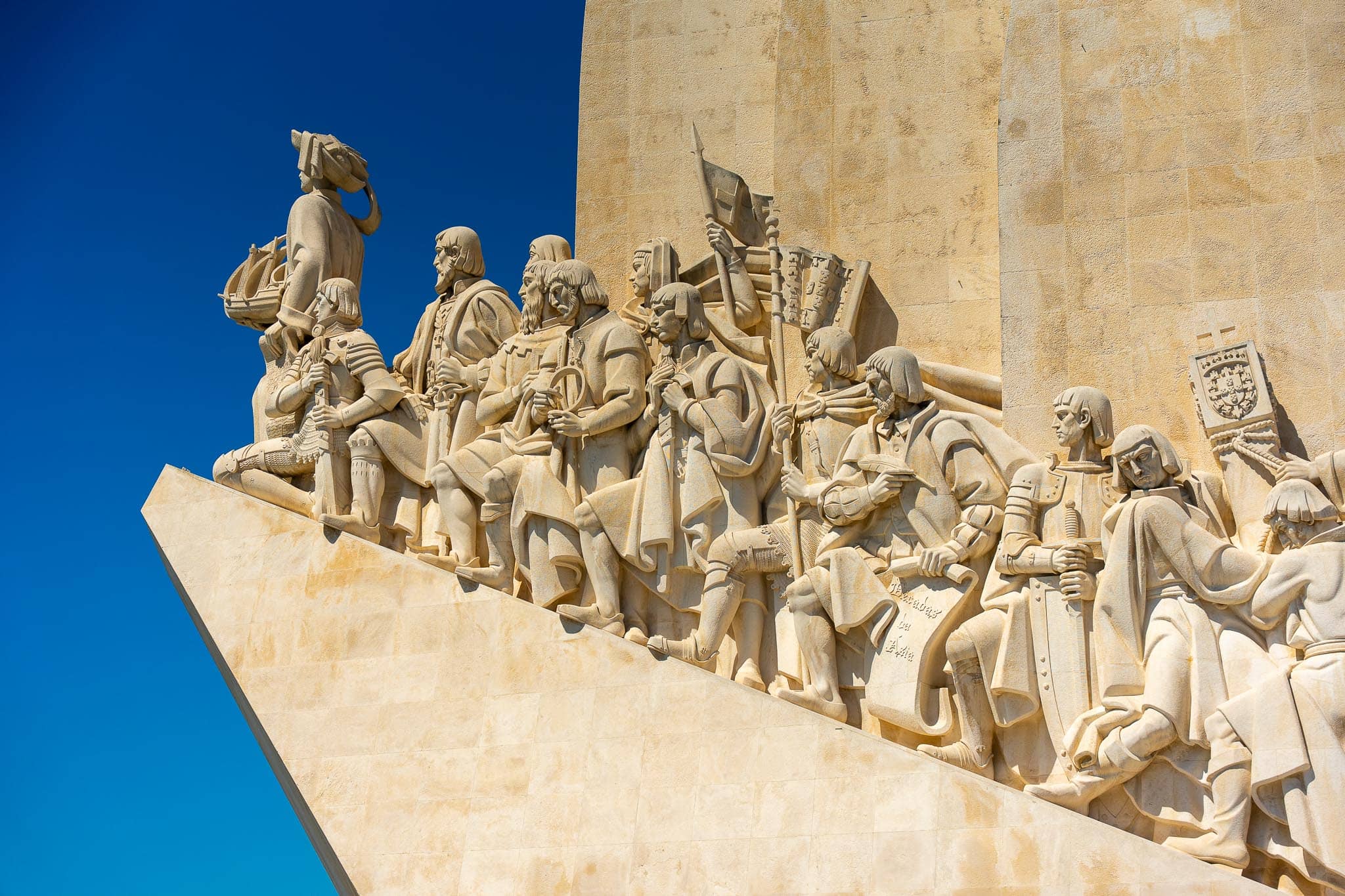 The carved stone prow of the Padrao dos Descobrimentos in Belem, a procession of navigators and clergy in pale limestone against a deep blue sky.