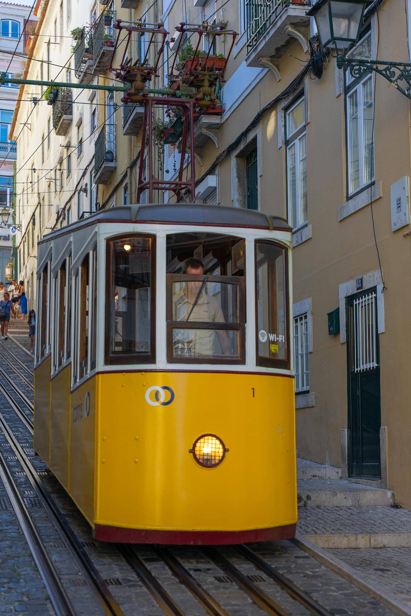 A yellow tram car standing on rails in a steep, narrow Lisbon street between pastel buildings with wrought-iron balconies.