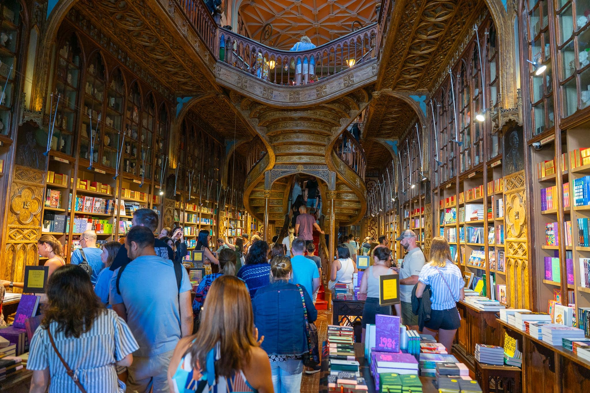 The interior of Livraria Lello in Porto, with carved wooden galleries, a stained-glass ceiling and a crimson staircase, packed with visitors browsing books.